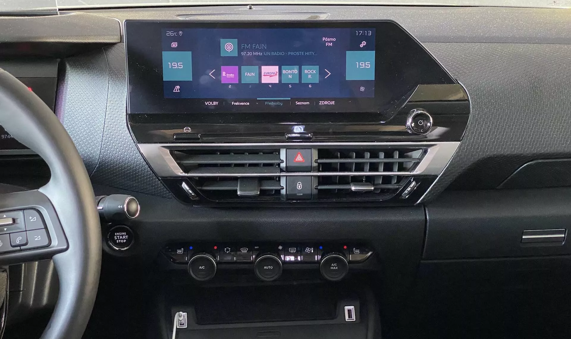 Pogled v notranjost Citroen e-C4 2020, ki je osredotočen na armaturno ploščo iz osrednjega položaja. Slika prikazuje sodoben zaslon na dotik z možnosti navigacije, medije, telefonsko povezljivostjo in nastavitvami avtomobila. Spodaj so horizontalni zračniki s gumbi za klimatsko napravo in druge funkcije, sledijo klimatski kontrolniki z dvema območjema s temperaturnimi gumbi in LCD zaslonom. Videti je del volana z vgrajenimi kontrolniki in gumb start-stop, kar poudarja sodobno avtomobilsko tehnologijo in ergonomsko zasnovo.
