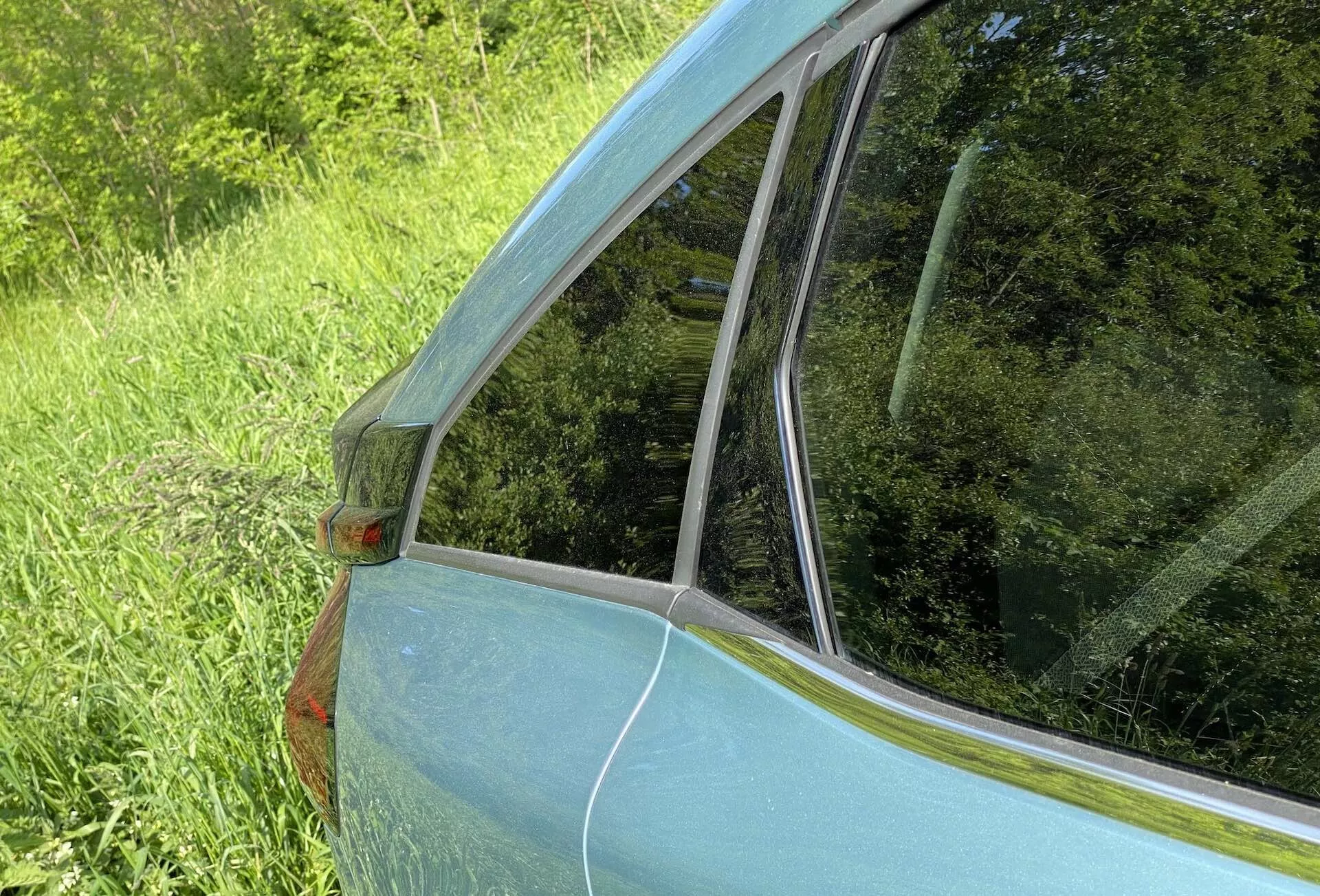 Podroben pogled na zadnji stranski del in okno Citroena C4 2020 (e-C4). Posnetek, narejen pod kotom od zadnje strani vozila, poudarja prehod od zadnjega potniškega okna do zadnjega vetrobranskega stekla. Vidni so kovinsko svetlo modra barva, oranžna stranska označevalna luč na stebru C, kromirana obroba in odsevi na steklu, kar poudarja dizajn in stanje avtomobila.