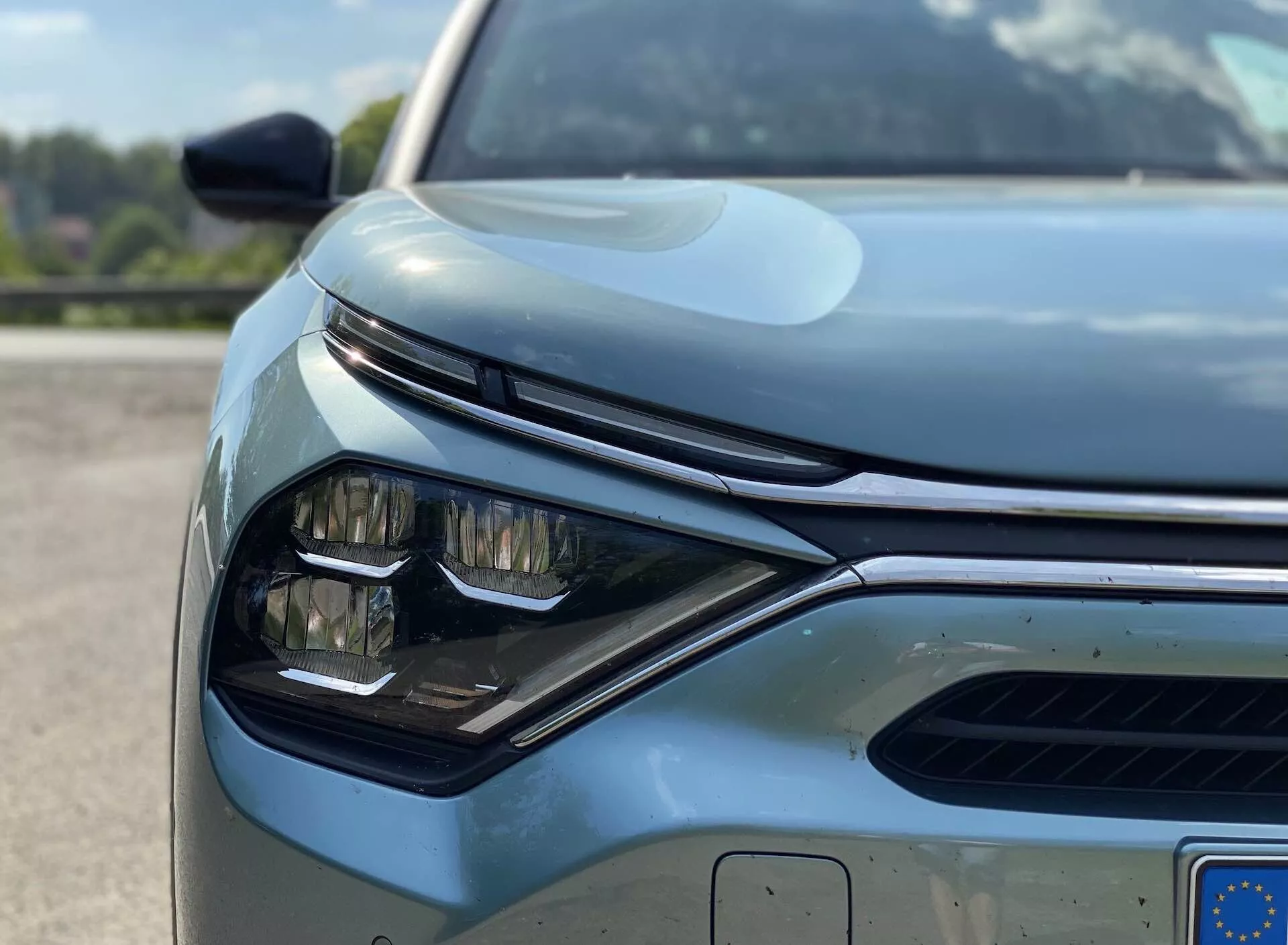 Pogled od blizu, pod nizkim kotom, na prednjo levo četrtino sodobnega vozila, specifično Citroen C4 2020 (e-C4). Osredotočeno na eleganten, kotni dizajn žarometa z vidnimi LED elementi, del mreže z vodoravnimi letvami, ki verjetno vključuje aktivni sistem loput, ter parkirne senzorje na sprednjem odbijaču. Slika zajema dele stranskega ogledala, opremljenega z indikatorji ali senzorji za nadzor mrtvih kotov. Pokrov motorja ima gladke linije z dvignjenim osrednjim delom, kar vozilu prinaša subtilen športni videz. Dobro vzdrževana, odsevna površina barve nakazuje visoko kakovost materiala ali učinkovito zaščitno prevleko. Delno vidna evropska registrska tablica poudarja sodobno avtomobilsko tehnologijo in oblikovanje.