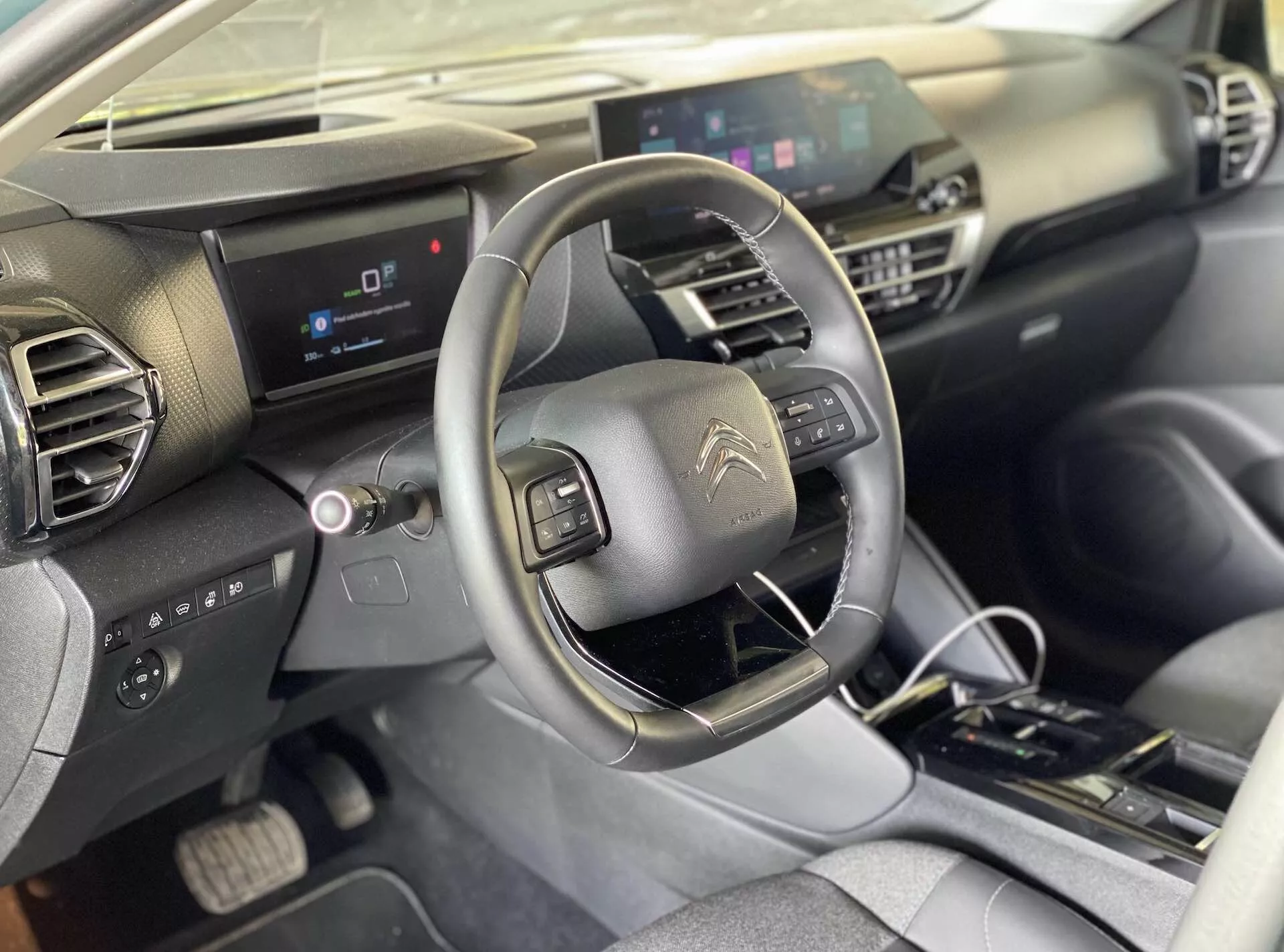 Pogled v notranjost Citroëna e-C4 2020 z vidika sprednjega sedeža sopotnika, ki zajema sedež voznika, osrednjo konzolo, volan z vgrajenimi kontrolami, digitalno instrumentno ploščo in osrednji zaslon informacijsko-zabavnega sistema. Armaturna plošča ima geometrijsko obliko s temnimi in svetlimi materiali. Osrednja konzola vključuje fizične kontrole za klimatsko napravo in avtomatski menjalnik.