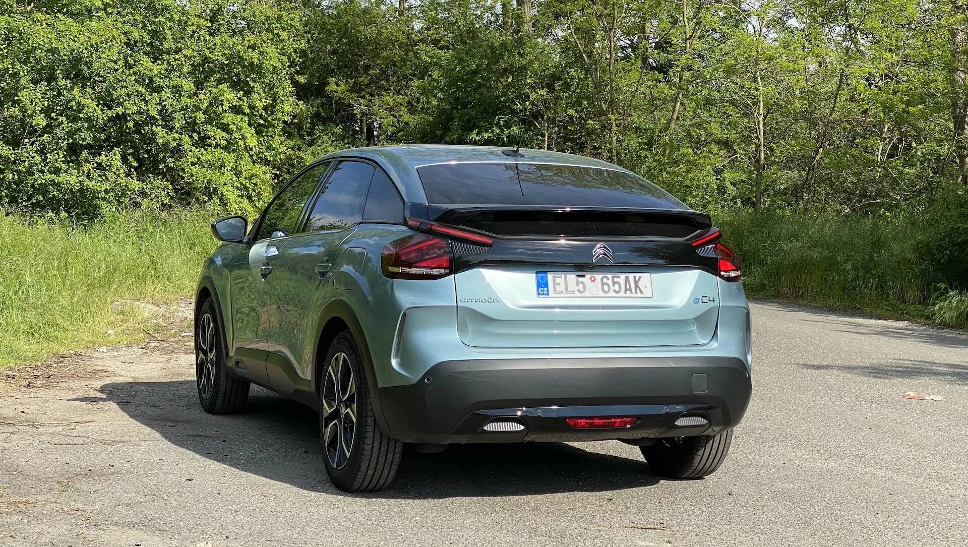 Zadnji kot parkiranega Citroena C4 2020 (e-C4) na asfaltni površini z naravno listjem v ozadju, ki prikazuje njegove značilne LED zadnje luči, oznake znamke in modela, zadnjo šipo z brisalcem, dvobarvni odbijač z elementom, ki je videti kot difuzor, vgrajene parkirne senzorje, delno vidni izpušni sistem, strešno anteno, platišča iz zlitin z več naperami, kovinski premaz barve in evropsko registrsko tablico.