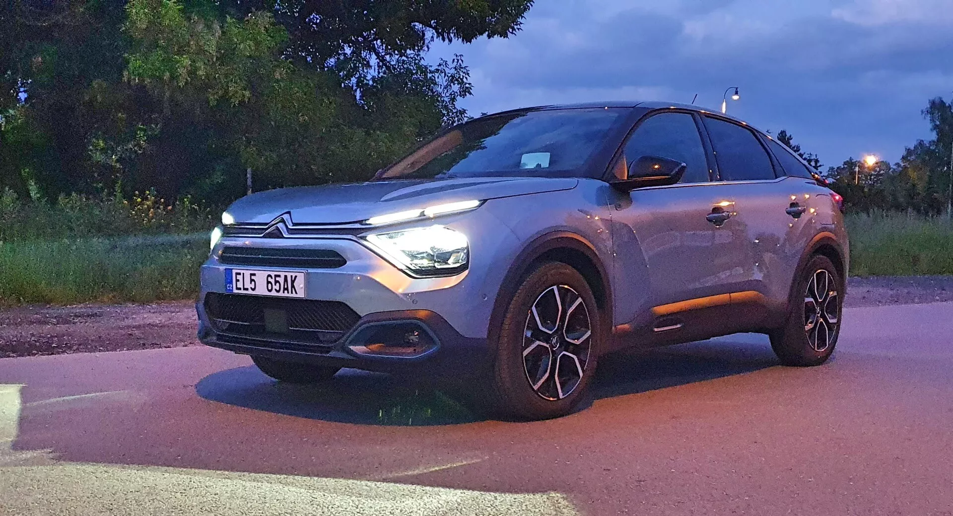 Citroen C4 (e-C4) 2020, moderni crossover SUV prikazan v mraku z osvetljenimi žarometi in dnevnimi lučmi. S tridimenzionalnim pogledom spredaj, slika prikazuje sprednjo fasado, mrežo z emblemom blagovne znamke, pokrov motorja, sprednji odbijač in bočni profil voznika, vključno z velikimi, večkrakimi litimi platišči. Vozilo, ki zaseda znaten del okvirja, prikazuje LED žaromete, značilno mrežo, ki bi lahko vključevala senzorje, ter kovinski lak, kar nakazuje višje ravni opreme ali možnosti prilagoditve.