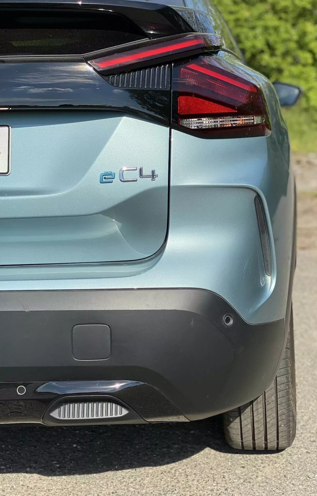 Zadnji kotni pogled na Citroën e-C4 2020, ki prikazuje značilni rdeči LED zadnji svetlobni sklop, ki se zdi, da se združuje z zadnjim vetrobranskim steklom, parkirni senzorji na odbijaču, del zadnjega difuzorja, značko modela 'e-C4', stilsko aluminijasto platišče in kovinsko barvo. Slika se osredotoča na zadnjo desno četrtino avtomobila iz rahlo dvignjenega in stranskega kota.