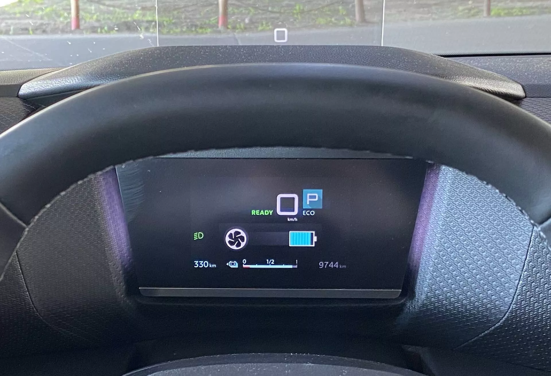 Od blizu prikaz iz perspektive voznika Citroëna C4 2020 (e-C4), ki prikazuje črno teksturiran volan in digitalno instrumentno ploščo. Volan rahlo zakriva spodnji del digitalnega zaslona, ​​ki prikazuje več funkcij: ikona pripravljenosti EV 'READY,' 'P' za parkiranje, način 'ECO,' skoraj popolnoma napolnjena ikona baterije, indikator dosega, ki prikazuje '330,' števec kilometrov '9744,' temperatura zunaj '+12°C' in ikona krožne puščice, verjetno za nastavitev moči regenerativnega zaviranja.