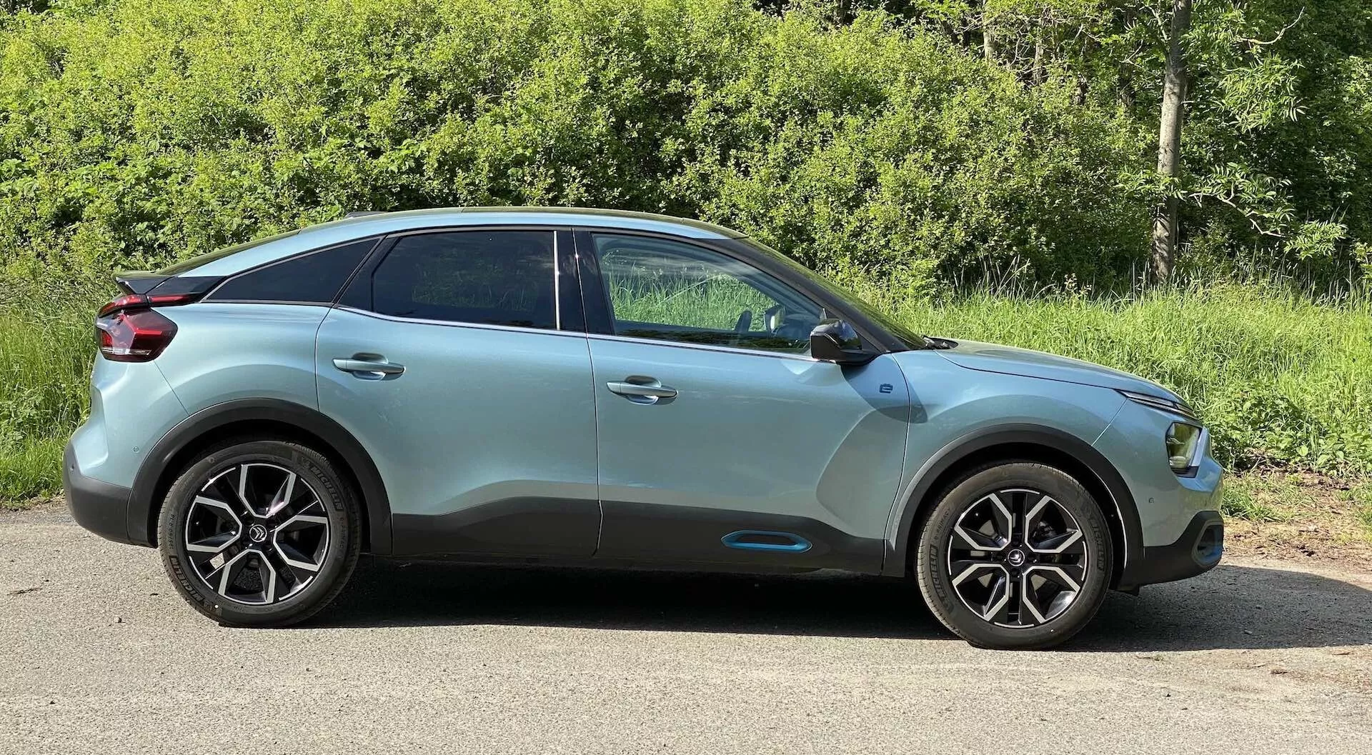 Stranski profil Citroen C4 (e-C4) 2020, prikazuje stran voznika iz rahlo nagnjenega prednjega perspektivnega kota. Slika poudarja moderne oblikovne linije vozila, aluminijasta platišča z značilno večkrako obliko, stranska ogledala v barvi karoserije z vgrajenimi smerniki in zatemnjena stekla na zadnjih oknih. Streha se nagiba proti zadnjemu delu, kar daje kupejevski videz telesu crossover/SUV. Avto ima kovinsko barvno končno obdelavo z vidnimi obrisi in oblikovnimi linijami, parkiran na ravni površini na prostem s ozadjem zelenega listja, osvetljen z naravno dnevno svetlobo.
