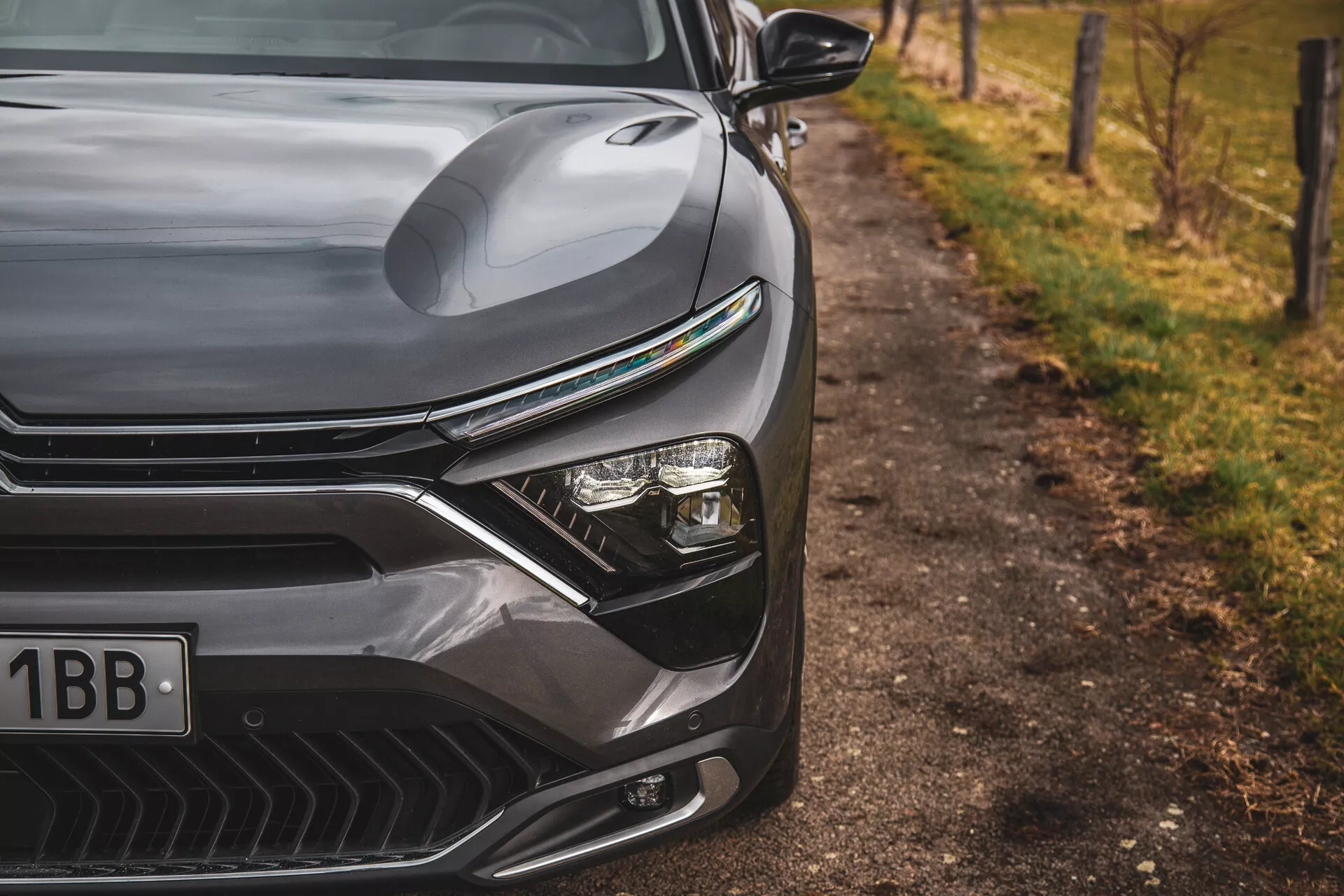 Posnetek iz bližine Citroena C5-X 2022, ki poudarja eleganten dizajn sprednje leve strani, vključno z oblikovanim pokrovom motorja, izrazito rešetko z horizontalnimi črtami, naprednim sistemom LED žarometov in aerodinamično oblikovanim sprednjim odbijačem z zračnimi vhodi, proti zamegljenemu naravnemu ozadju ob cesti.