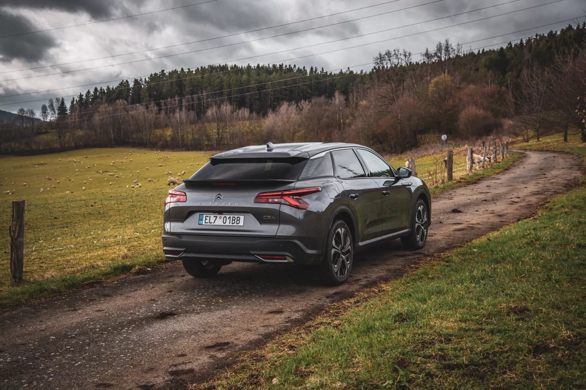 Citroen C5-X 2022, sodoben hatchback parkiran na nepokriti podeželski cesti, zadnji tričetrtinski pogled razkazuje aerodinamično zasnovo z zadnjim spojlerjem in morsko-plavutno anteno, vitke vodoravne zadnje luči in velika kolesa z večkrakim dizajnom, ki nakazujejo zmogljivost in luksuz v nenujanem okolju.