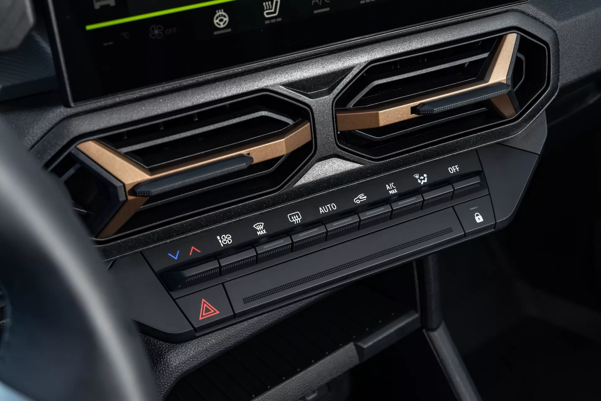 Pogled od blizu na sistem za klimatsko kontrolo v Dacia Duster 3. generacije 2024 na centralni armaturi pod zaslonom za informacije in zabavo, ki prikazuje nastavitve temperature in hitrosti ventilatorja, funkcije razdeljevanja zraka, lastnosti odtaljevanja, gumb za vključitev opozorilnih luči in možnosti za klimatizacijo, obdano z kotnimi zračniki in poudarjeno z metalik obrobo za sodobno estetiko notranjosti