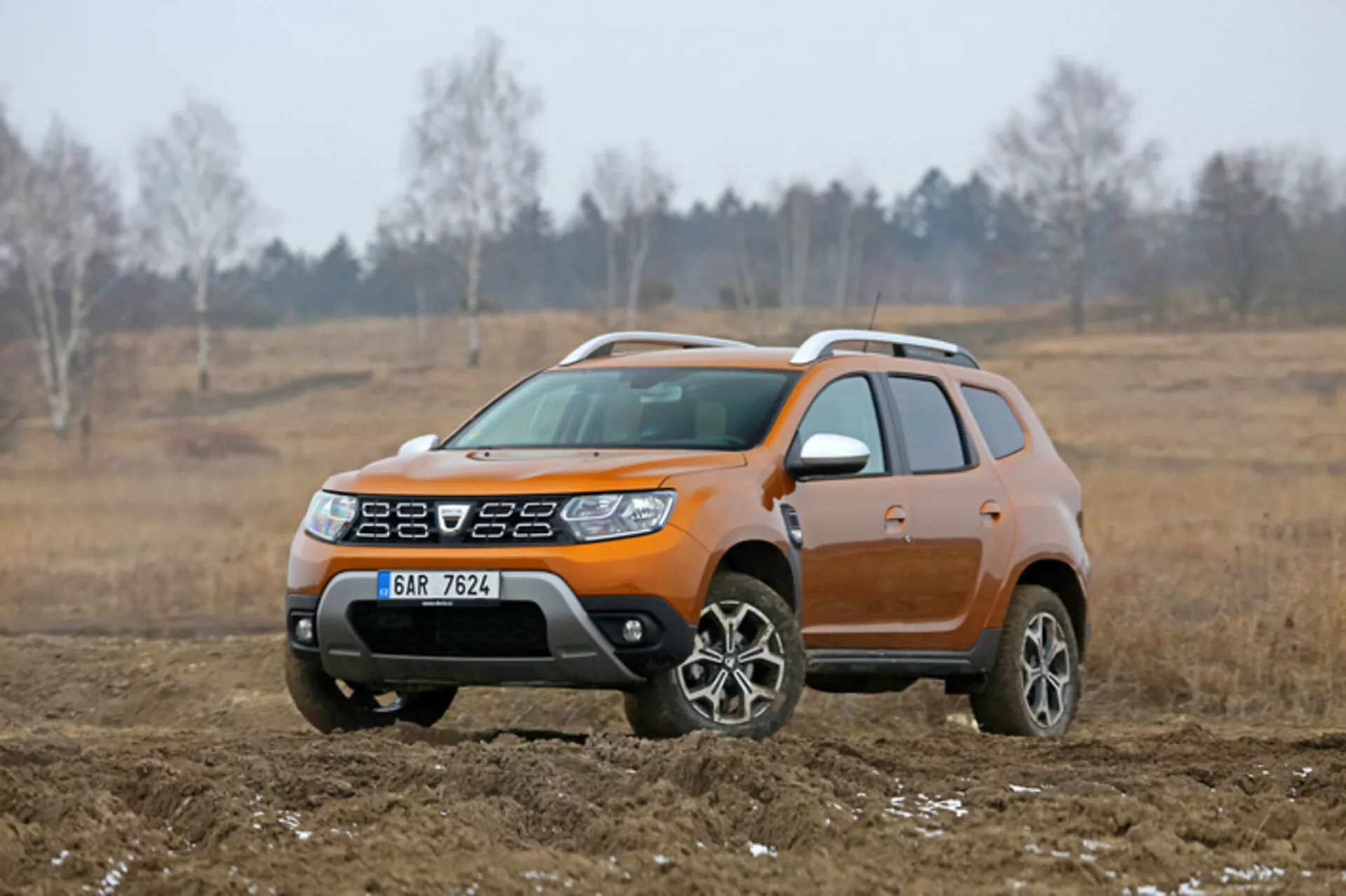 Kompaktni SUV Dacia Duster iz leta 2013, predstavljen v zunanjem, terenskem okolju, poudarjen z njegovo značilno oranžno barvo, dvignjeno višino vožnje, lita platišča in plastičnim oplatom telesa za lahko terensko uporabo, ujet iz kota tri četrtine spredaj.
