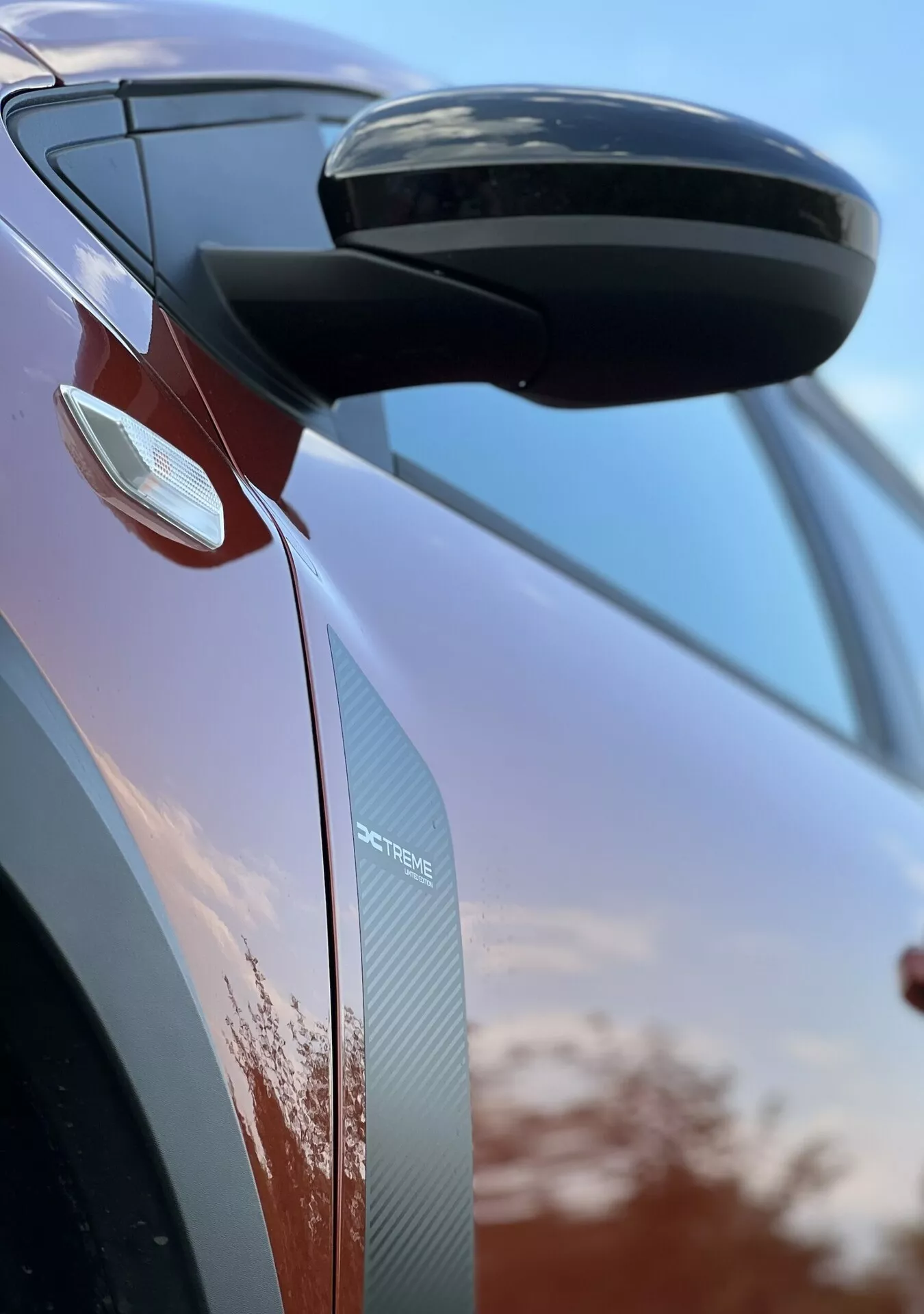 Podrobni pogled na Dacia Jogger 2022, osredotočen na bočno vzvratno ogledalo in oblikovanje vrat, ki prikazuje aerodinamične značilnosti, integrirane smernike in teksture iz ogljikovih vlaken na ozadju neba