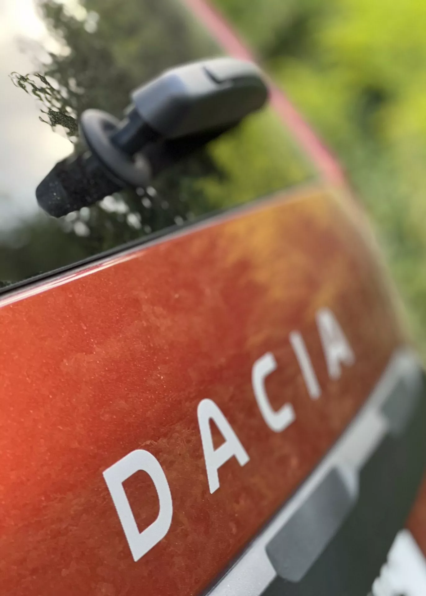 Pogled od blizu na zadnji panel Dacia Jogger 2022 z napisom DACIA z velikimi belimi črkami na metalik oranžno-rdeči barvi z madeži in umazanijo, ki nakazuje zunanjo uporabo in vzdržljivost, postavljen v naravnem okolju z zamegljenim stranskim ogledalom, ki nakazuje sodobne lastnosti ogledala.