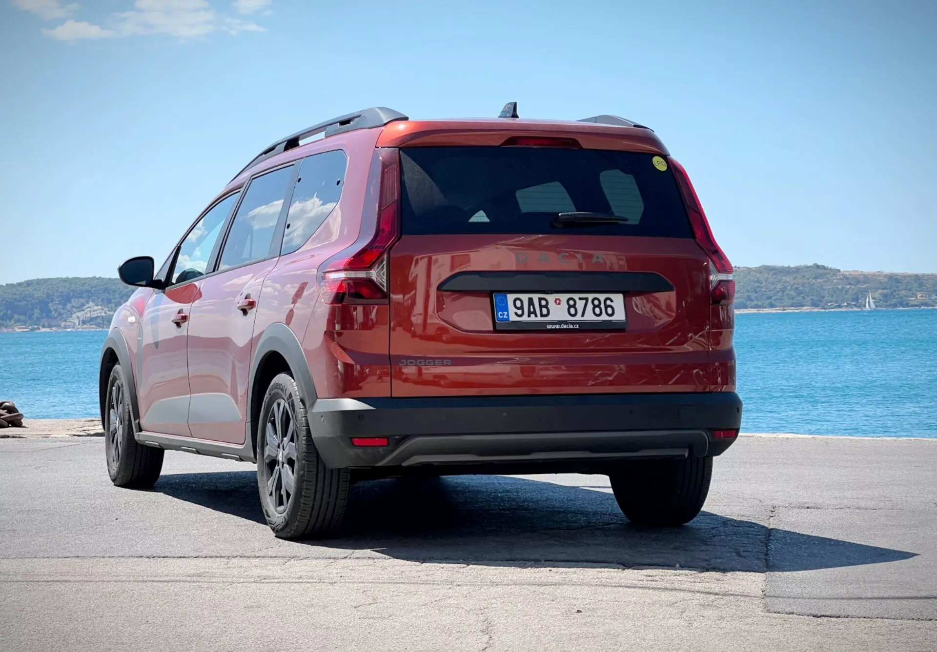 Dacia Jogger 2022 v rdeči barvi, posneta iz zadnjega kota treh četrtin, ki prikazuje stranico in obliko prtljažnih vrat SUV, parkirana v bližini vodne površine z obzorjem v ozadju, poudarjajoč njena platišča iz lahkih zlitin z večimi kraki, LED zadnje luči in aerodinamični zadnji spojler