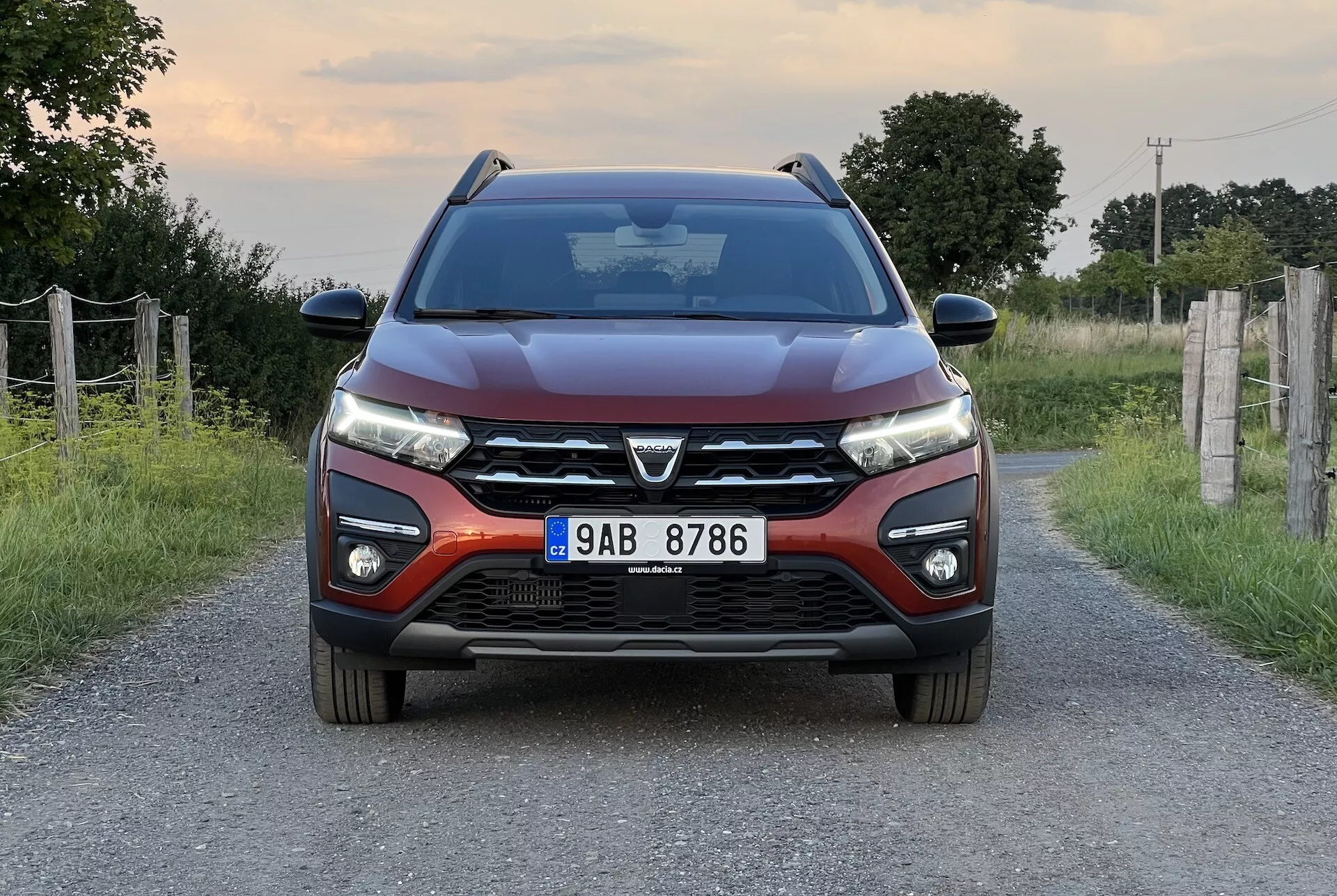 Dacia Jogger 2022, pozicioniran na ozki podeželski cesti, prikazuje simetričen sprednji pogled, ki poudarja moderno obliko avtomobila, vključno z izstopajočo sprednjo masko z velikim emblemom, elegantnimi LED žarometi, oblikovnimi linijami pokrova motorja, sprednjim odbijačem z meglenkami in parkirnimi senzorji, ter stranskimi ogledali z integriranimi obratomerkami.