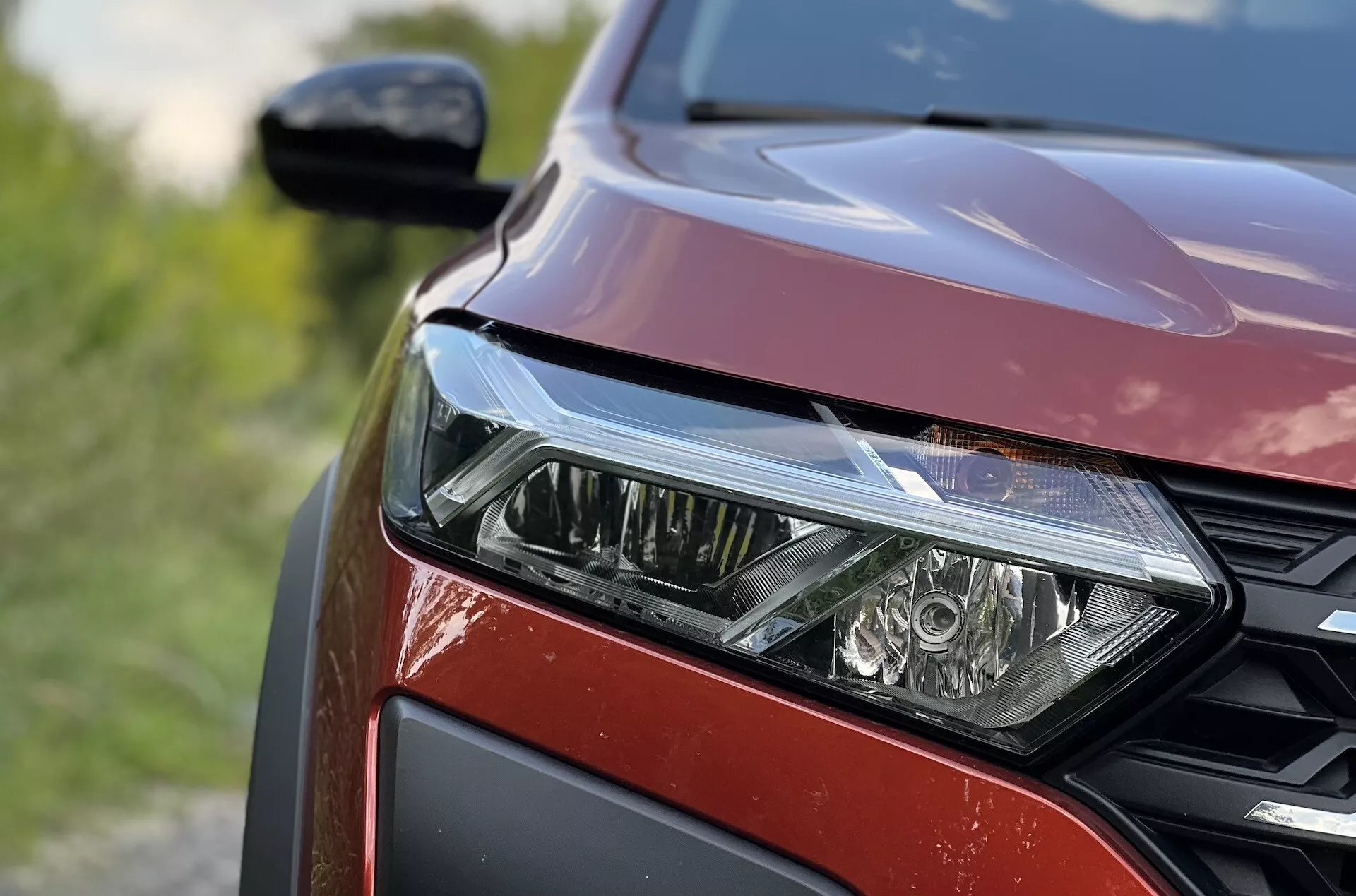 Podroben pogled na sprednji levi del Dacia Jogger 2022, ki poudarja sklop LED žarometov, vzorec satovja na rešetki in kovinsko rdečo barvo laka