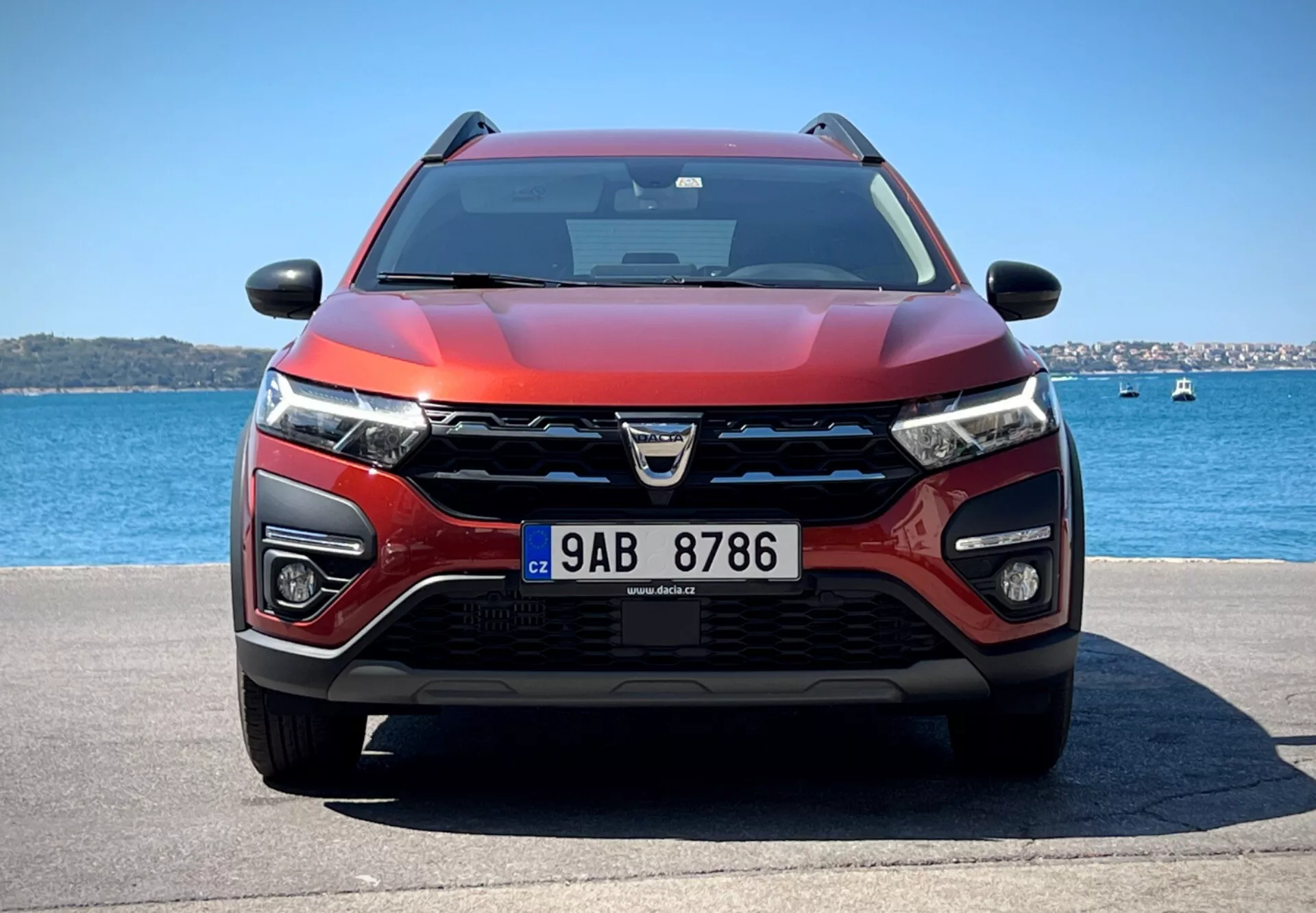 Dacia Jogger 2022 predstavlja svoj sodoben crossover dizajn in napredne tehnološke lastnosti v mirnem vodnem okolju, z izrazito rešetko z logotipom, LED žarometi, meglenkami, potencialnimi senzorji ADAS, dovodi zraka za hlajenje in radar, dvobarvno rdeče in črno barvno shemo, aerodinamičnim pokrovom, opremljen z brisalci, ki nakazujejo možno tehnologijo zaznavanja dežja, stranskimi ogledali z integriranimi smerniki, morda vsebujejo tehnologijo za spremljanje mrtvega kota, pod jasnim nebom.