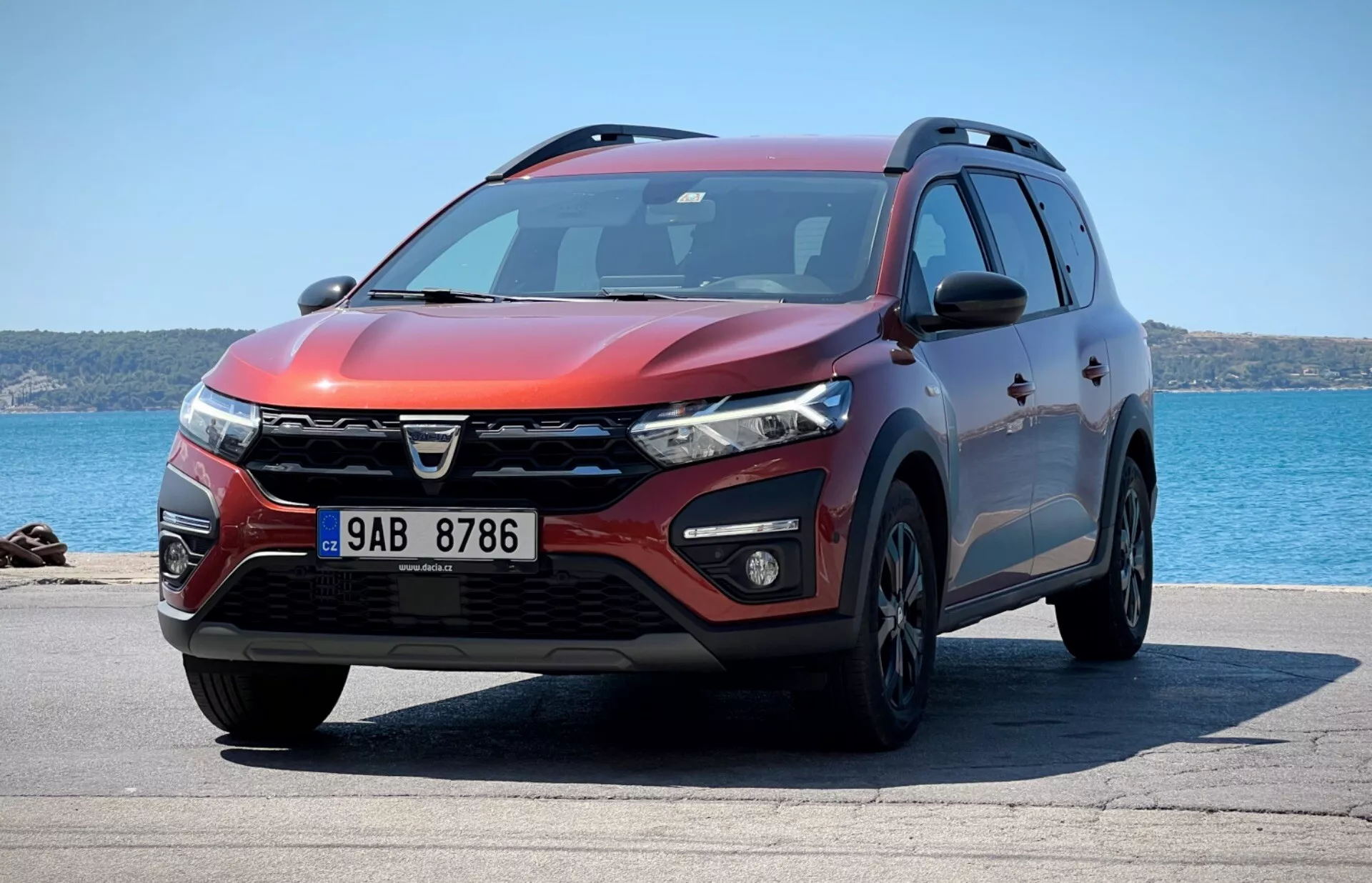 Vid tri četrt spredaj del SUV Dacia Jogger 2022, parkiranega z ozadjem oceana, prikazuje značilno obliko rešetke, LED žaromete, meglenke, aerodinamični sprednji odbijač z zračnimi vstopi in parkirnimi senzorji, mišičaste konture pokrova, zlitinska kolesa, integrirane smerne kazalnike na stranskih ogledalih, stranski profil s zaščitno oblogo, strešne nosilce, zatemnjena okna in metalik rdečo zunanjo barvo pod svetlim sončnim svetlom, kar kaže na oblikovni profil, tehnološke značilnosti in uporabnost.