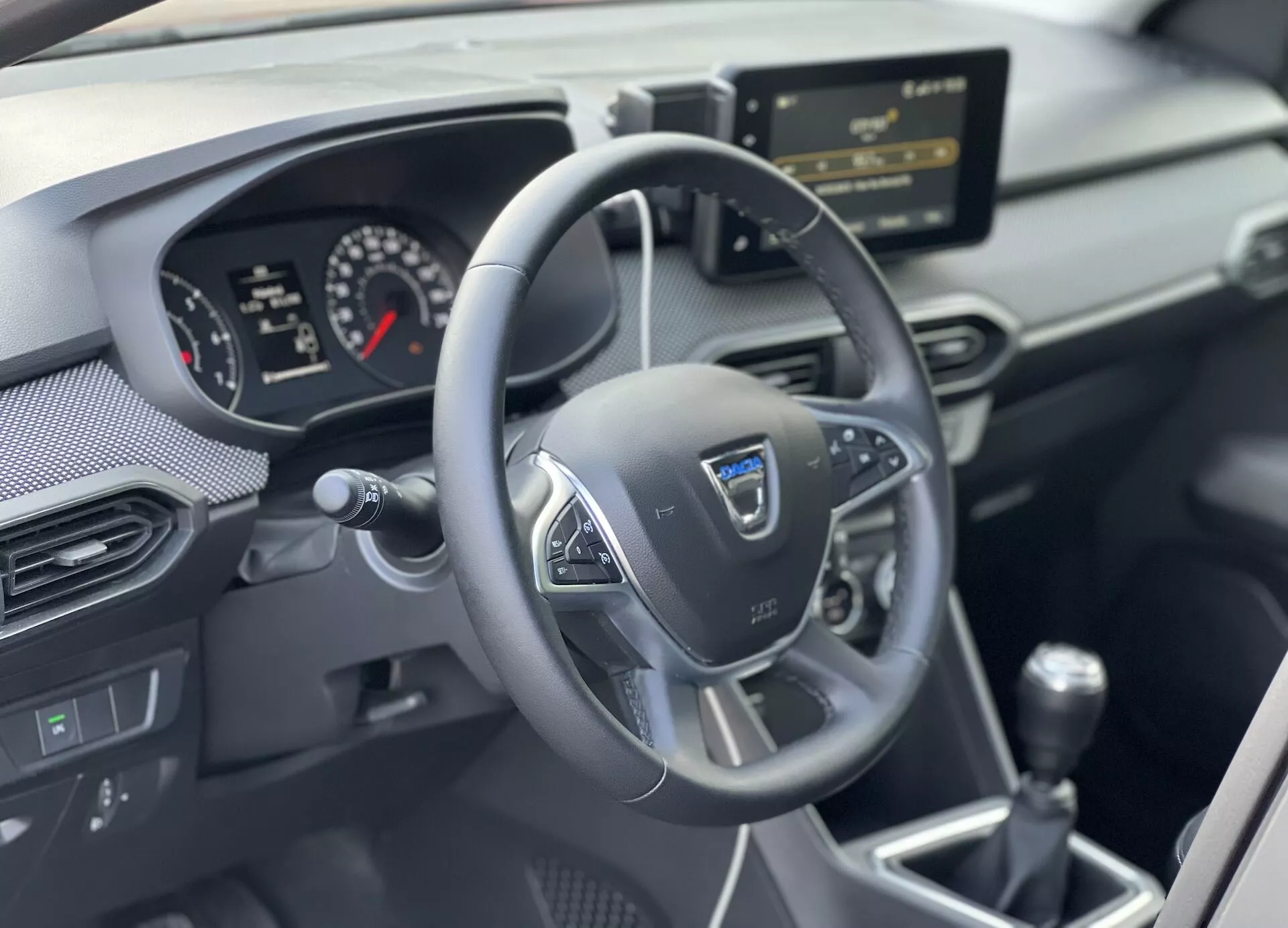 Notranjost Dacia Jogger 2022 osredotočena na voznikovo področje s trirakim multifunkcijskim volanom, analognim-digitalnim instrumentnim sklopom, sredinsko konzolo z infotainment sistemom in ročico ročnega menjalnika