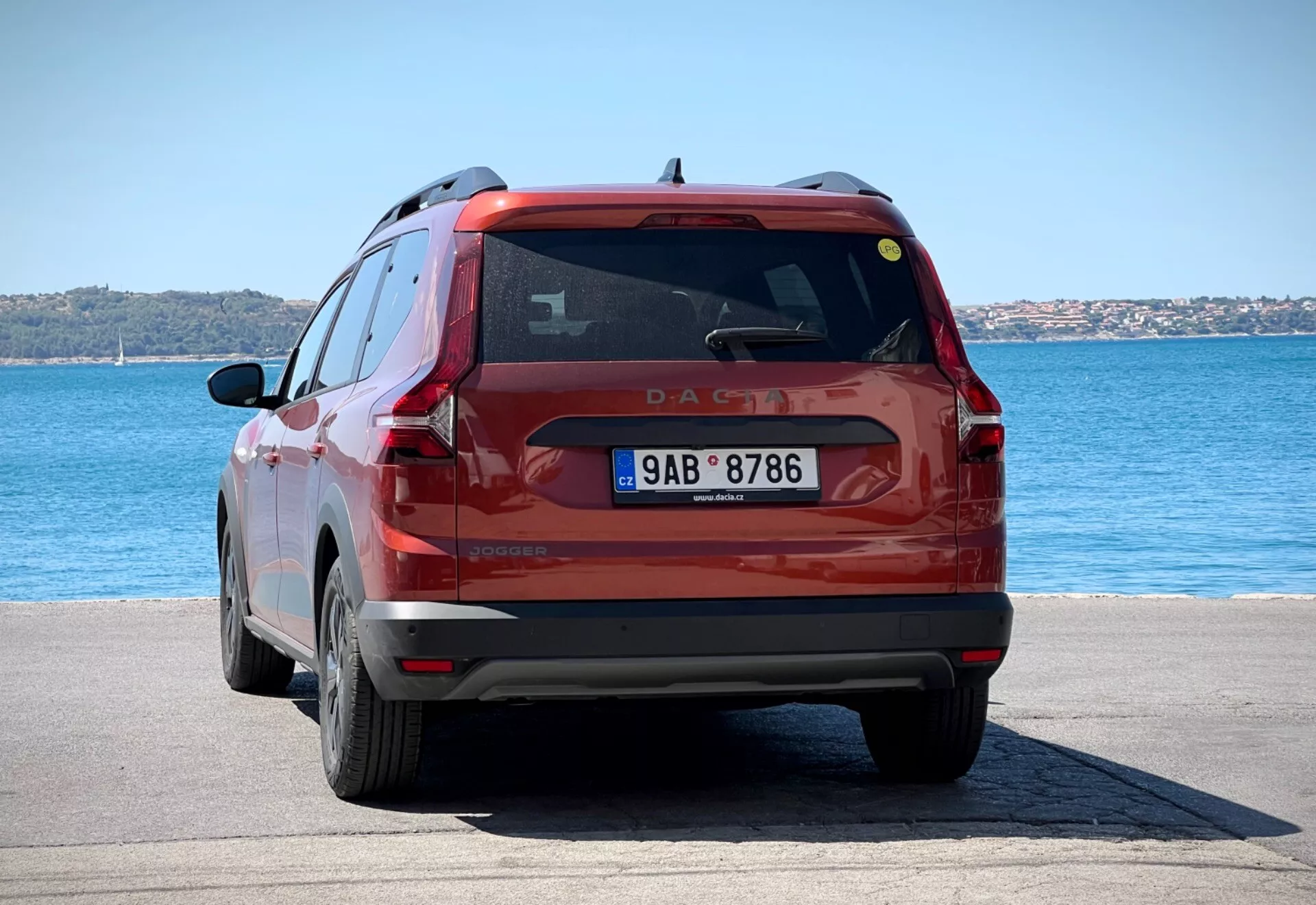 Zadnji pogled na rdeč kompaktni SUV Dacia Jogger 2022, parkiran na tlakovanem obrežju pod svetlo dnevno svetlobo, prikazuje celotni zadnji del vozila, vključno z zadnjimi lučmi, zadnjim vetrobranskim steklom in registrsko tablico proti ozadju modre vode in jasnega neba.