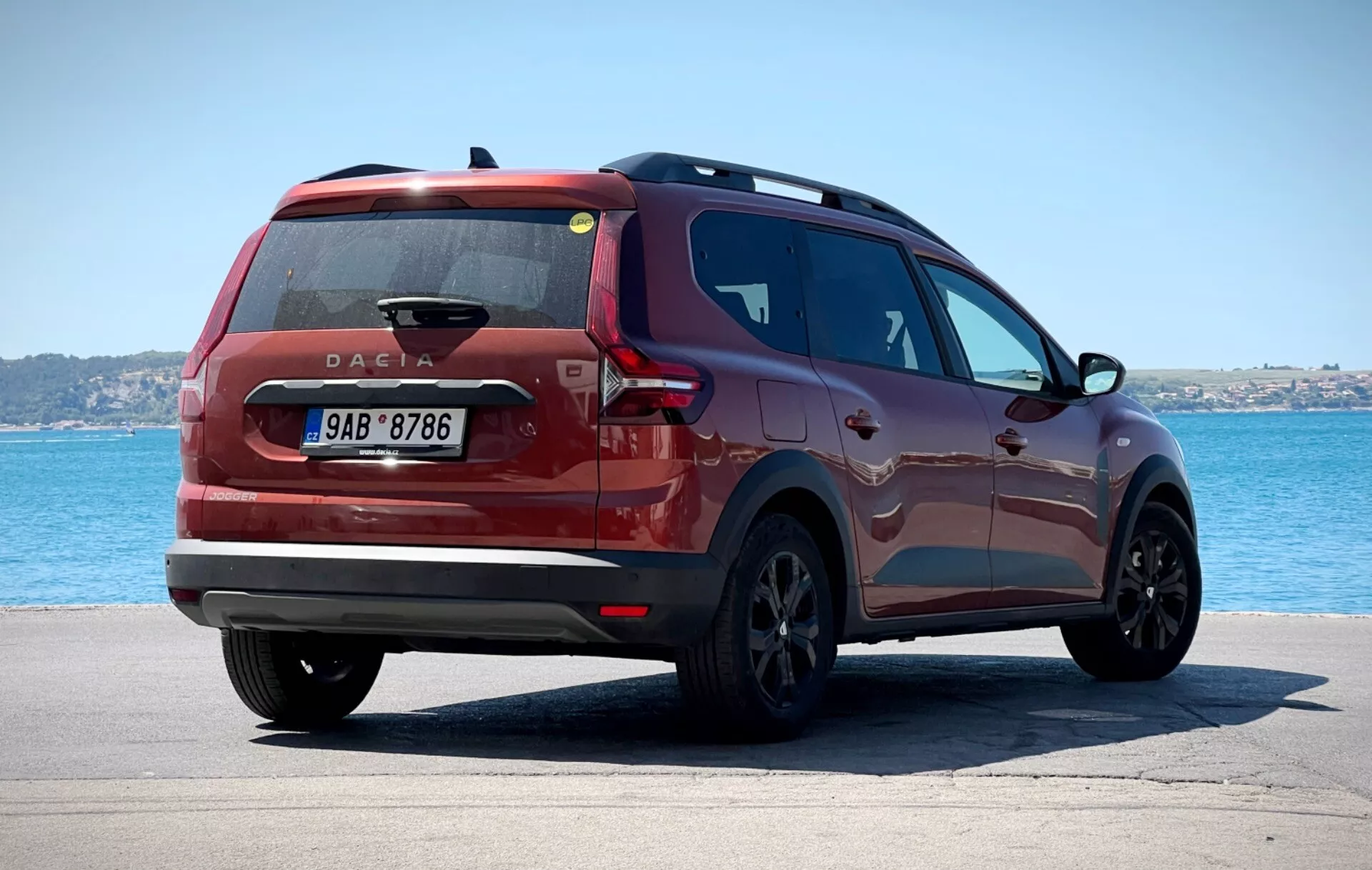 Dacia Jogger 2022 fotografirana iz tričetrtinskega zadnjega pogleda, postavljena na tlakovani površini z vodno površino in jasnim nebom v ozadju, prikazuje moderne zadnje luči, centralno postavljeno značko Dacie in eleganten bočni profil proti mirnemu obalnemu ali jezerskemu okolju.