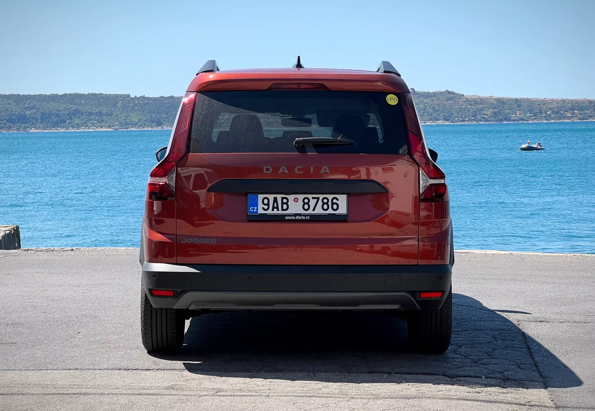 Zadnji pogled na rdeč Dacia Jogger 2022, ki prikazuje prtljažna vrata z emblemo in imenom modela, zadnje vetrobransko steklo z brisalcem in tretjo zavorno lučjo, moderne zadnje luči, registrsko tablico in odbijač z reflektorji in potencialnimi senzorji za parkiranje, parkiran ob obali ob dnevu.
