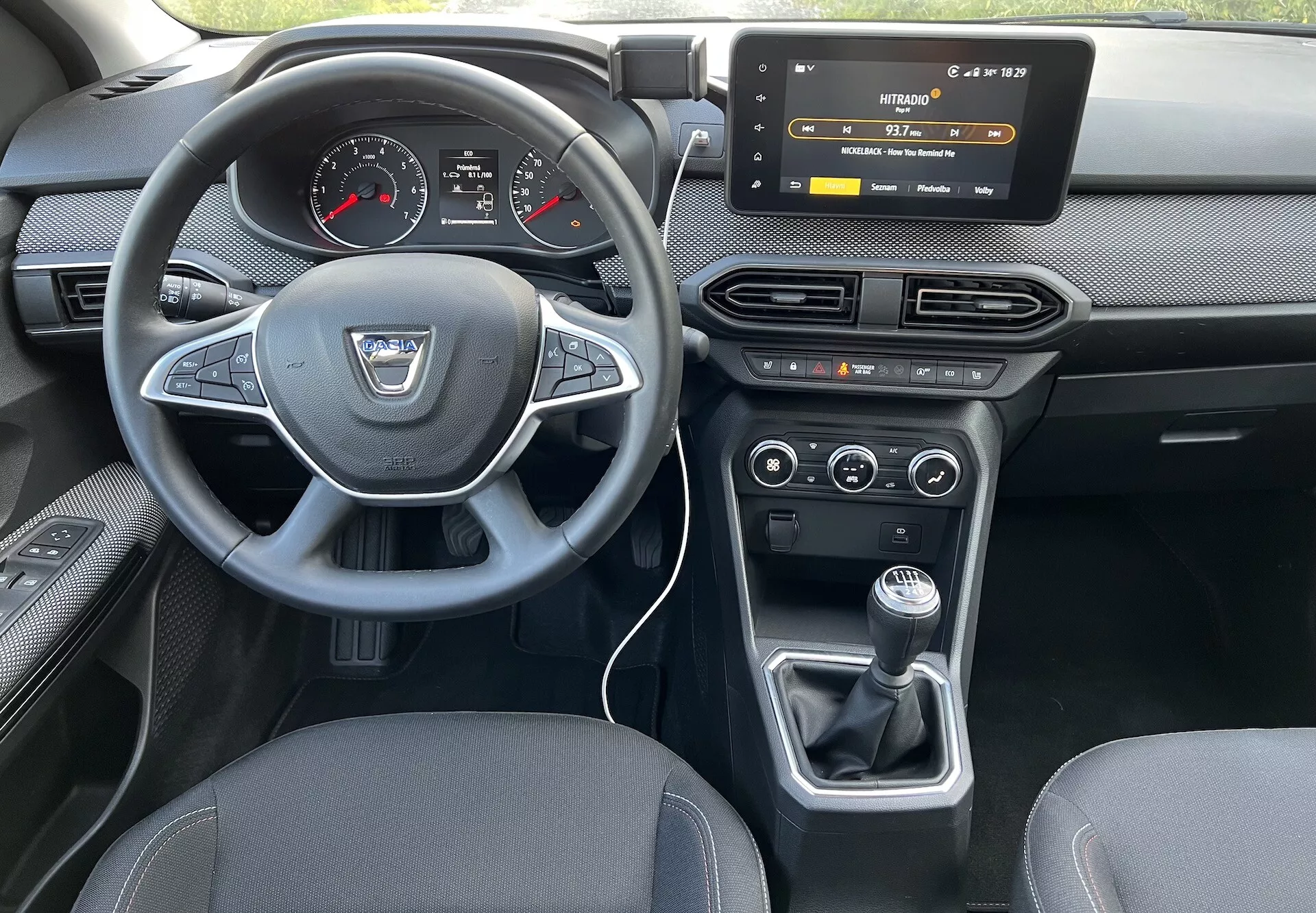 Notranji pogled Dacia Jogger 2022 iz zadnjega sedeža, ki poudarja armaturno ploščo voznika, volan z logotipom proizvajalca in kontrolnimi gumbi, digitalno-analogni instrumentni sklop, zaslon na dotik infotainment sistema, kontrole HVAC, klimatsko napravo z dvojnim območjem in ročni prestavni mehanizem