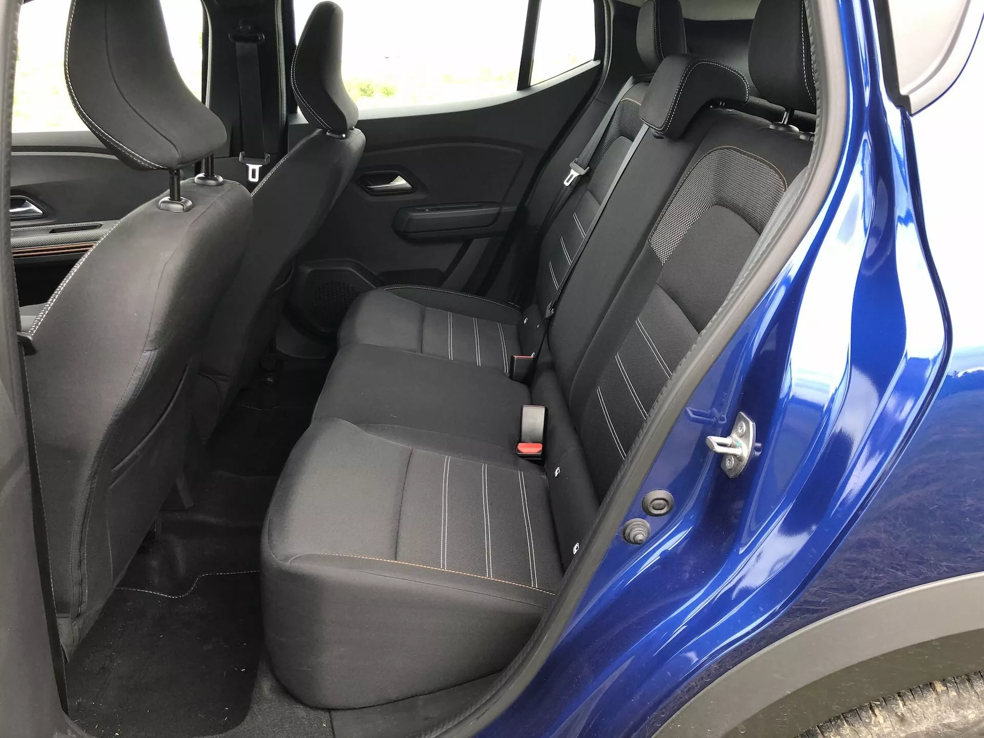 Notranjost Dacia Sandero-2020 (Sandero-Stepway) s poudarkom na zadnjem sedežnem prostoru za potnike. Posneta nekoliko nad ravnjo sedežev, prikazuje desno stran z varnostnimi pasovi, kljuko vrat in temno barvno shemo s črno plastiko in temno sivo ali črno oblazinjenje. Poudarja konfiguracijo sedežev, varnostne funkcije, kot so nastavljivi varnostni pasovi in možne pritrditvene točke ISOFIX, nadzor električnih oken na vratnem panelu in celotno zasnovo notranjosti.