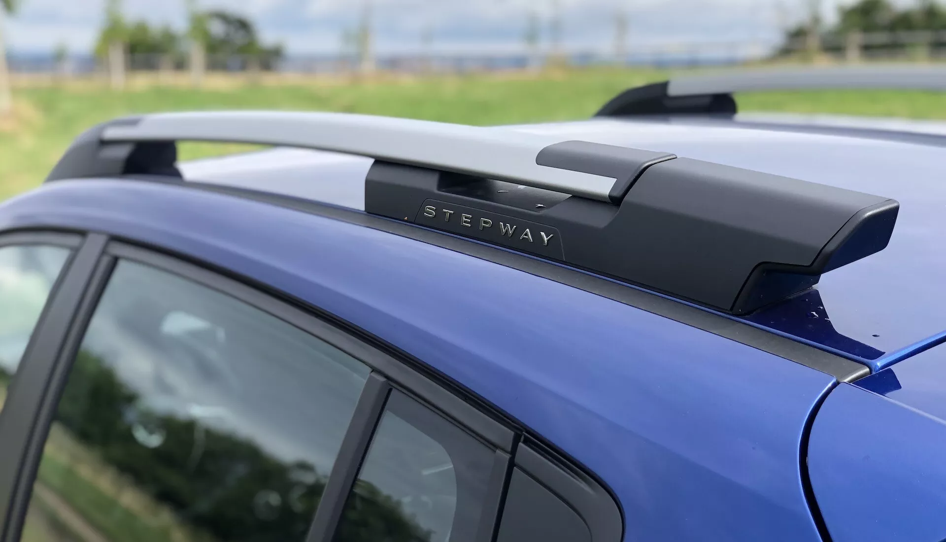 Bližnji pogled na zadnji del strehe modrega Dacia Sandero Stepway 2020, ki poudarja kovinske strešne nosilce z oznako 'STEPWAY', aerodinamično zasnovo strehe in sijajni barvni premaz, kar nakazuje dobro vzdrževano vozilo, zasnovano za dodatno shranjevanje tovora.