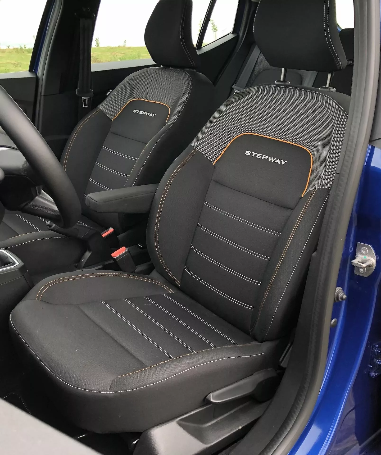 Podroben pogled na notranjost sprednjih sedežev vozila Dacia Sandero 2020 Stepway, prikazuje voznikov sedež z izrazitimi stranskimi ojačitvami za dodatno podporo in vezenje 'STEPWAY' v beli barvi z oranžnim obrisom. Sedeži imajo dvobarvno barvno shemo, prevladuje črna z teksturiranim vzorcem in vidnimi dvojnimi šivi. Fotografija zajema stranske profile sedežev iz rahlo dvignjenega kota, razkriva zgornji del in del sedežne površine, kar nakazuje na športno in vzdržljivo opremo. Vidni del interierja vključuje majhen del modrega zunanjega okvirja vrat.