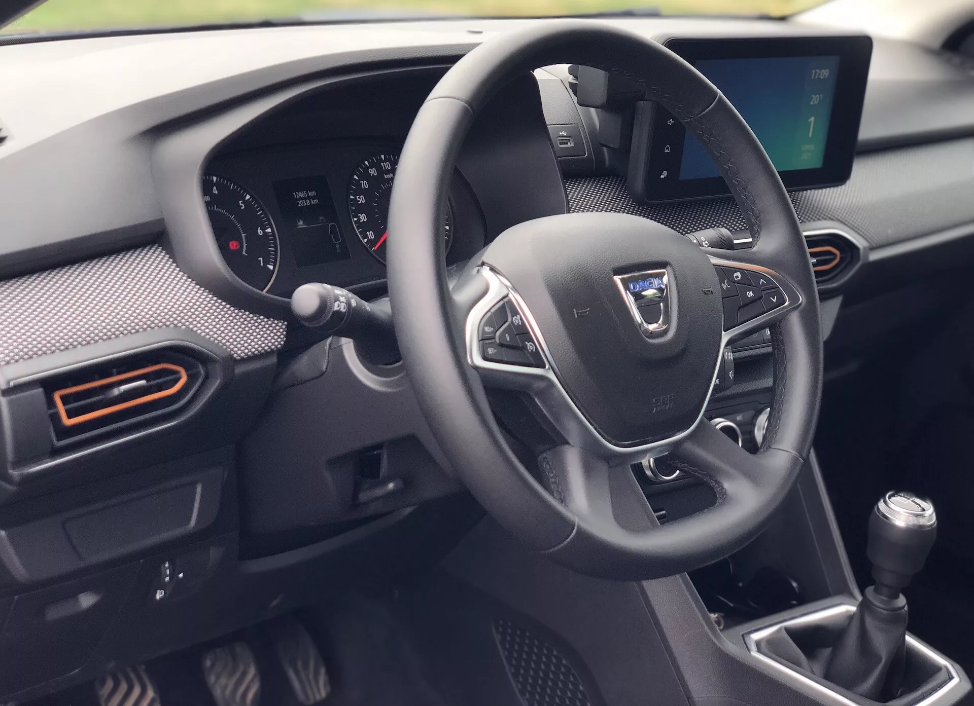 Notranjost vozila Dacia Sandero-Stepway 2020 z izpostavljenim trikrakam volanom z večpredstavnostnimi upravljalniki, delnim pogledom na armaturno ploščo z analognimi in digitalnimi prikazi, ročnim menjalnikom in podrobno sredinsko konzolo s sistemom za zagon na gumb. Slikana poudarja sodobno tehnologijo in športno oblikovanje z oranžnimi poudarki na pravokotnih zračnikih in elementih, ki spominjajo na karbonska vlakna.