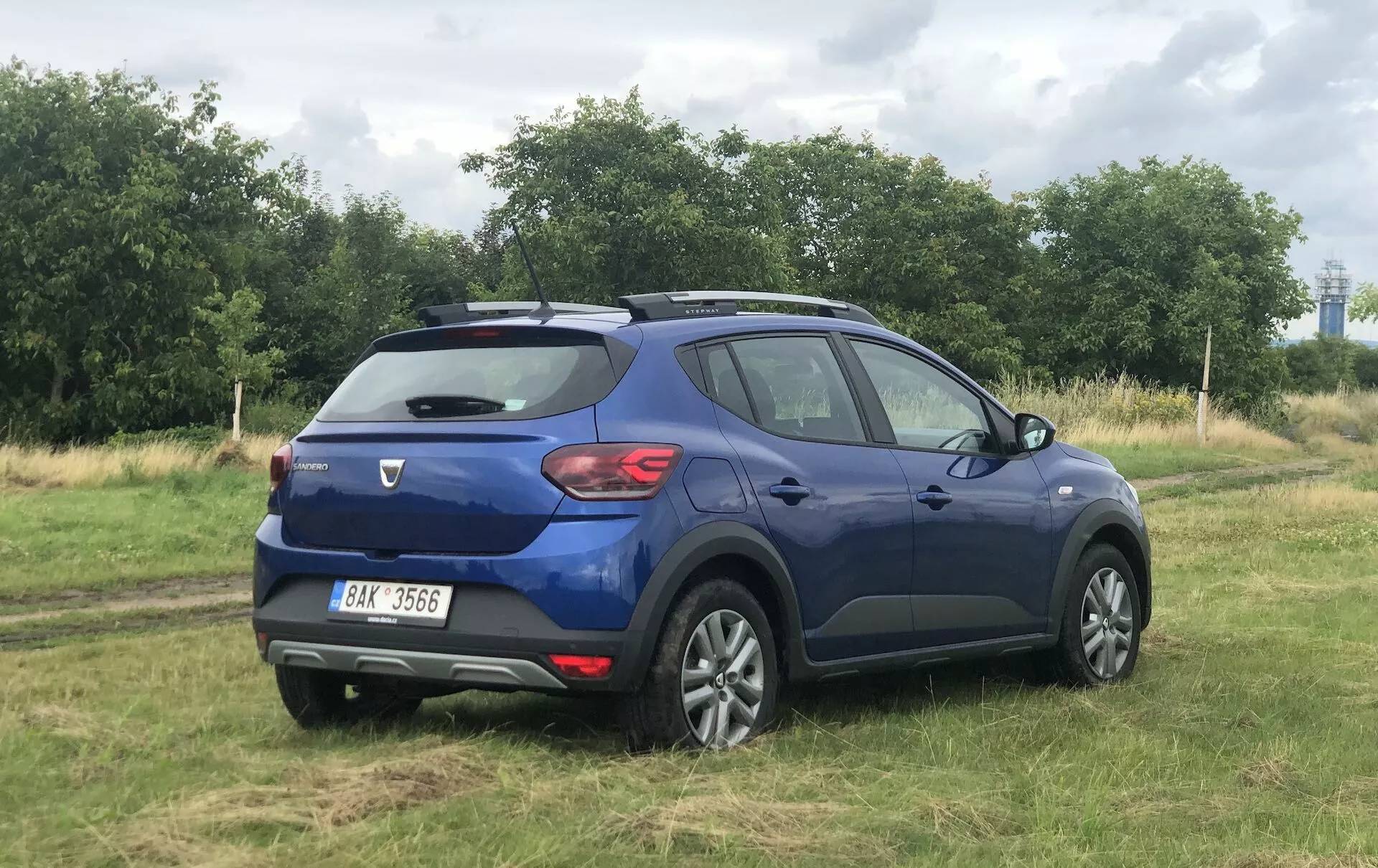 Ta slika prikazuje modri Dacia Sandero Stepway 2020, kompaktni crossover SUV, parkiran na travniku, posnet iz tričetrtinskega zadnjega pogleda. Poudarja zadnja vrata z brisalcem, zadnje luči, zadnji spojler, značke znamke in modela, srebrno zaščitno ploščo, dvobarvna lita platišča, strešne nosilce, zatemnjena okna ter linije in obrise karoserije vozila.