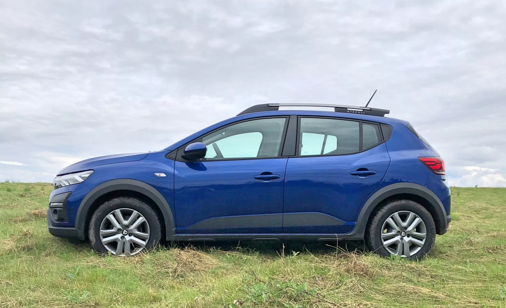 Slika modrega Dacia Sandero Stepway 2020, crossover SUV, parkiranega na odprtem polju. Avto je prikazan iz profila, poudarja sodoben dizajn sprednje luči, litkasta platišča in teksturirane pnevmatike. Zunanje lastnosti vključujejo stranska ogledala, kljuke vrat, karakterne linije in rahlo izbočenje nad blatniškimi loki. Strešna linija rahlo pada proti zadnjemu delu in vključuje strešni sistem, kar povečuje uporabnost. Vidimo tudi namig zadnje luči in zatemnjena stekla na zadnjih oknih. Ozadje prikazuje rahlo zamegljeno oblačno nebo in travo, zaradi česar je vozilo glavni predmet slike.