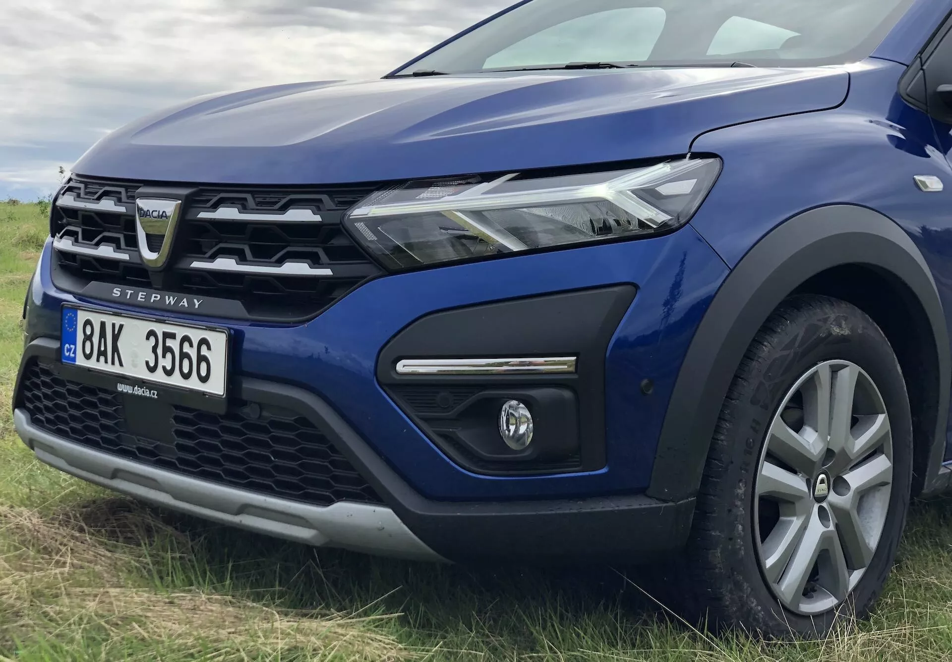 Od blizu fotografija leve prednje četrtine modrega Dacia Sandero Stepway 2020, ki prikazuje sodoben žaromet z LED osvetlitvijo, delno vidno rešetko z značilnim emblemom, prednji odbijač z meglenko v črnem okviru, dvignjen pokrov motorja, aluminijasto kolo z več naperami, stransko ogledalo v barvi karoserije, plastične obloge na blatniku in vratih, ki nakazujejo na terenske zmogljivosti, nagnjeno vetrobransko steklo in A-stebriček, ter oznako 'STEPWAY', ki označuje model.