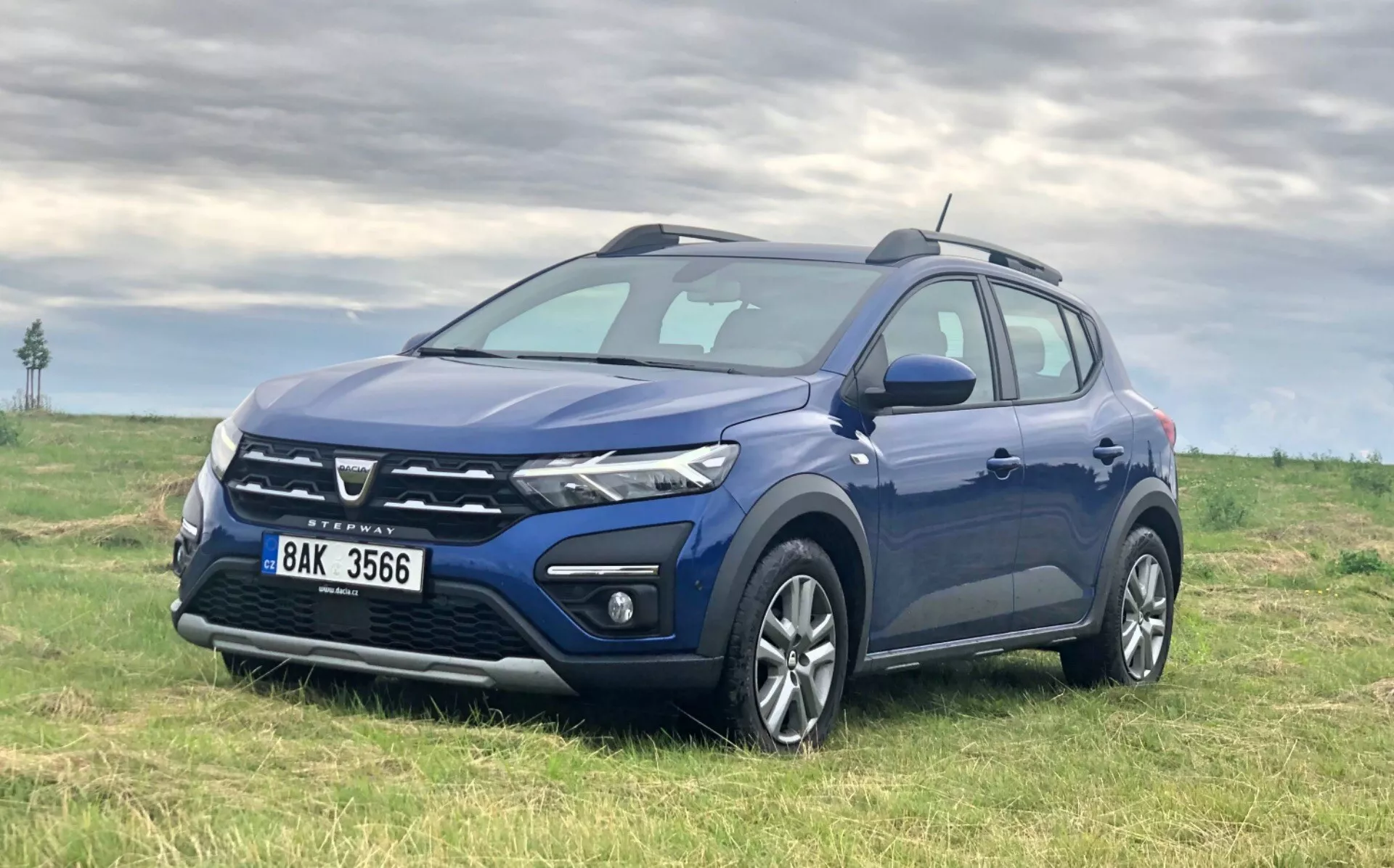 Slika temno modrega kompaktnega SUV-ja Dacia Sandero 2020 (Sandero Stepway) parkiranega na travnatem polju z rahlo oblačnim nebom v ozadju, prikazuje sprednjo rešetko, žaromete, sprednji odbijač, kolesa in profil na voznikovi strani iz tričetrtinskega sprednjega kota, rahlo dvignjeno.