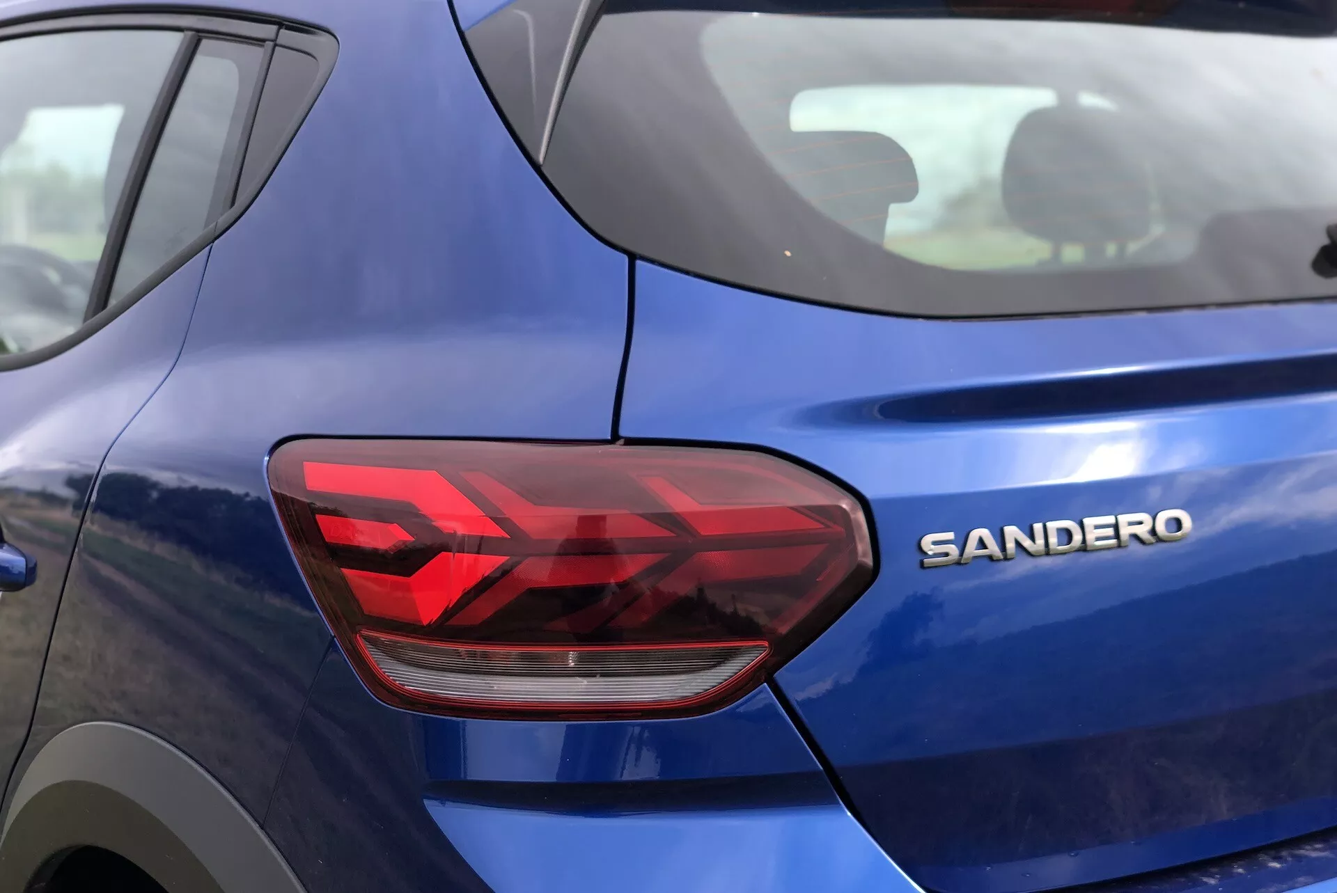 Bližnji bočni pogled na zadnji del modrega Dacia Sandero-Stepway 2020 z poudarkom na sodobnih kotnih LED zadnjih lučeh in kromiranem napisu 'SANDERO'. Slika prikazuje kovinsko barvno obdelavo, zadnjo luč s sekcijami za zavorne luči, smernike in vzvratne luči ter ostre, geometrijske oblikovne elemente, ki nakazujejo novi model hatchbacka ali majhnega avtomobila.