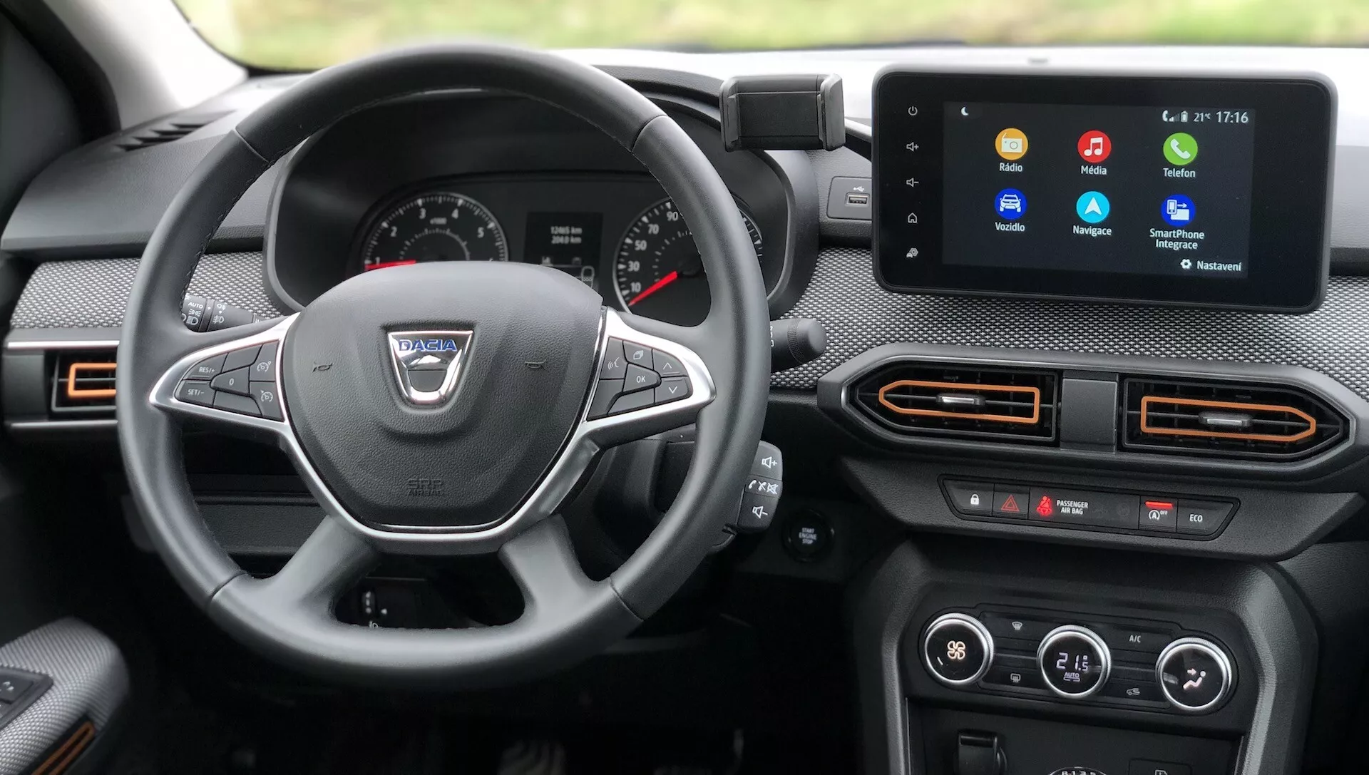 Notranji pogled iz voznikovega sedeža Dacia Sandero 2020 (Sandero Stepway), prikazujúc armaturno ploščo, volan in sredinsko konzolo. Armaturna plošča vsebuje mešanico analognih in digitalnih zaslonov z dvema velikima analognima številčnicama in manjšimi merilniki, vključno s centralnim digitalnim zaslonom. Moderna trikraka volanska kolesa z večfunkcijskimi gumbi poudarja infotainment sistem. Sredinska konzola vključuje zaslon na dotik z različnimi ikonami in zračniki z oranžnimi poudarki. Spodaj so fizični gumbi in vrtljivi gumbi za nadzor klime. Vidni materiali v kabini vključujejo kombinacijo tekstur, ambientalna osvetlitev pa je vidna v oranžni svetlobi okoli zračnikov.