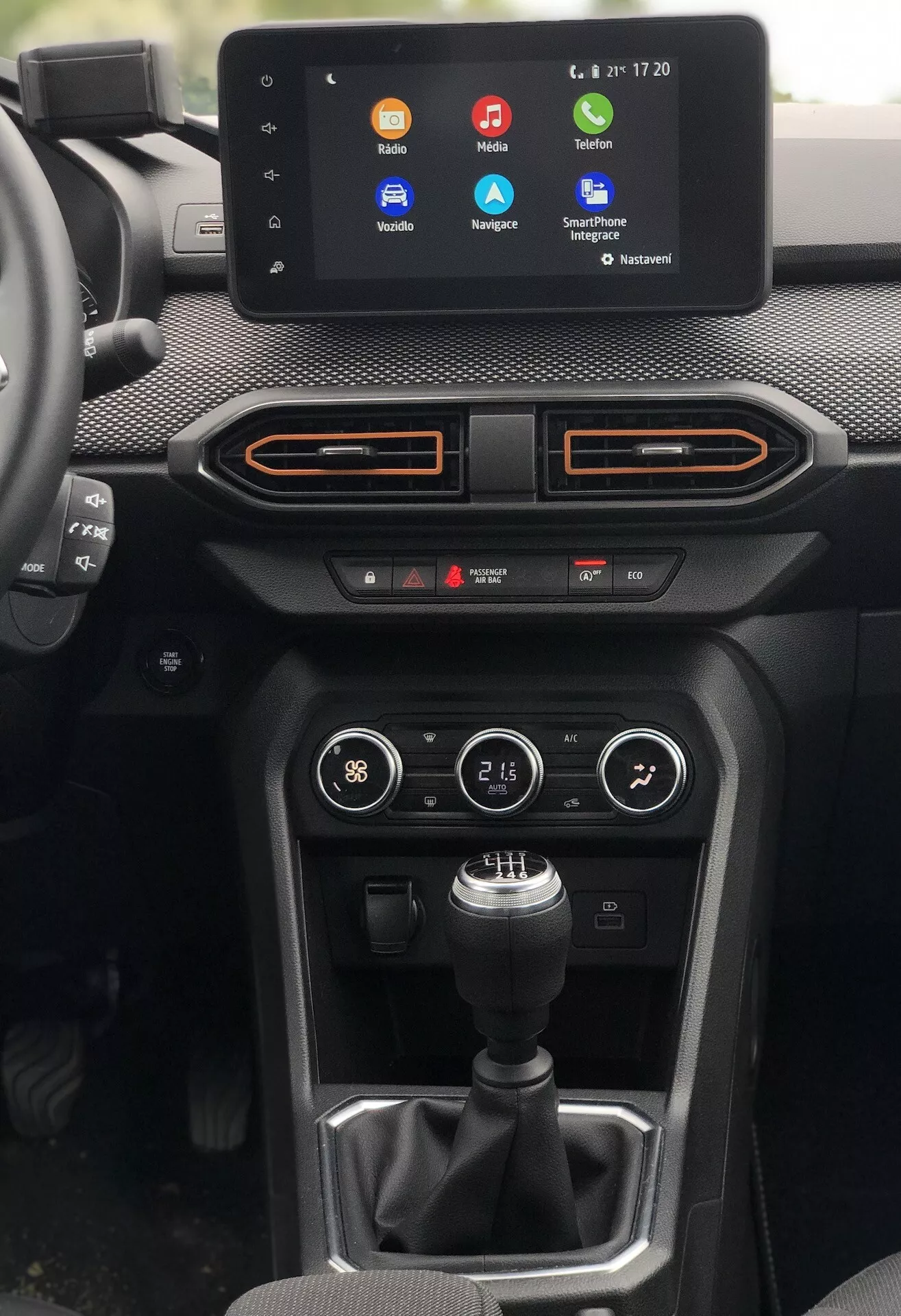 Notranji pogled na Dacia Sandero Stepway 2020, osredotočen na sredinsko konzolo z voznikove perspektive. Pomembne funkcije vključujejo informativni zaslon na dotik, ki prikazuje pisane aplikacijske ikone za klice, navigacijo in pretakanje glasbe, s časom in temperaturo v zgornjem desnem kotu. Pod zaslonom sta dve vodoravni zračni reži z oranžnimi poudarki, plošča za nadzor klime z gumbi in rotacijskimi gumbi ter ročica ročnega menjalnika na teksturiranem ozadju. Gumb na desni strani nakazuje različne vozne načine ali kontrole, takoj pod kontrolami klime pa sta vidna dva USB vhoda. Uporabljeni materiali so mešanica mehke armaturne plošče na dotik, sintetične ali tekstilne teksture in plastike. Poleg tega je vzdolž robov zračnih rež ambientalna osvetlitev, ki se ujema z oranžnim poudarkom. Sestava ponuja jasen pogled brez popačenja, rahlo nagnjena v desno za uravnotežen prikaz.