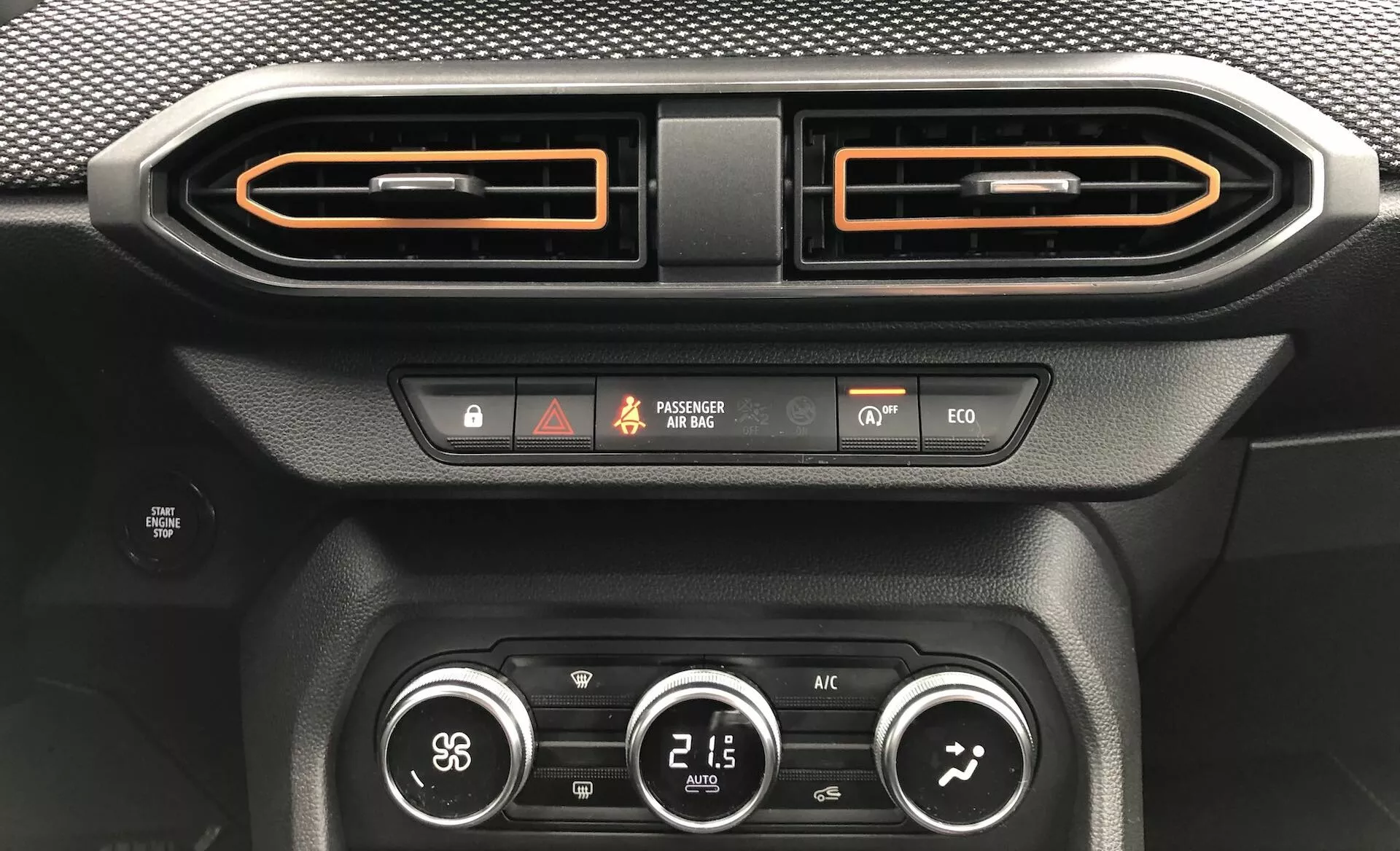 Pogled od blizu na notranjost avtomobila Dacia Sandero 2020 (Sandero-Stepway), osredotočen na območje osrednje armature. Slika prikazuje dve vodoravni odprtini za zrak s kovinskim zaključkom in oranžno obrobo, gumb za opozorilne luči, indikator stanja zračne blazine za sovoznika, indikator varnosti vozila, gumb za način ekološke vožnje, gumb za odmrzovanje in klimatski sistem s tremi rotacijskimi gumbi, ki prikazujejo hitrost ventilatorja, temperaturo (21,5 °C) in smer pretoka zraka. Drugi detajli vključujejo teksturo, podobno ogljikovim vlaknom, in mat črne materiale, osvetljen prikaz in osvetljene gumbe.