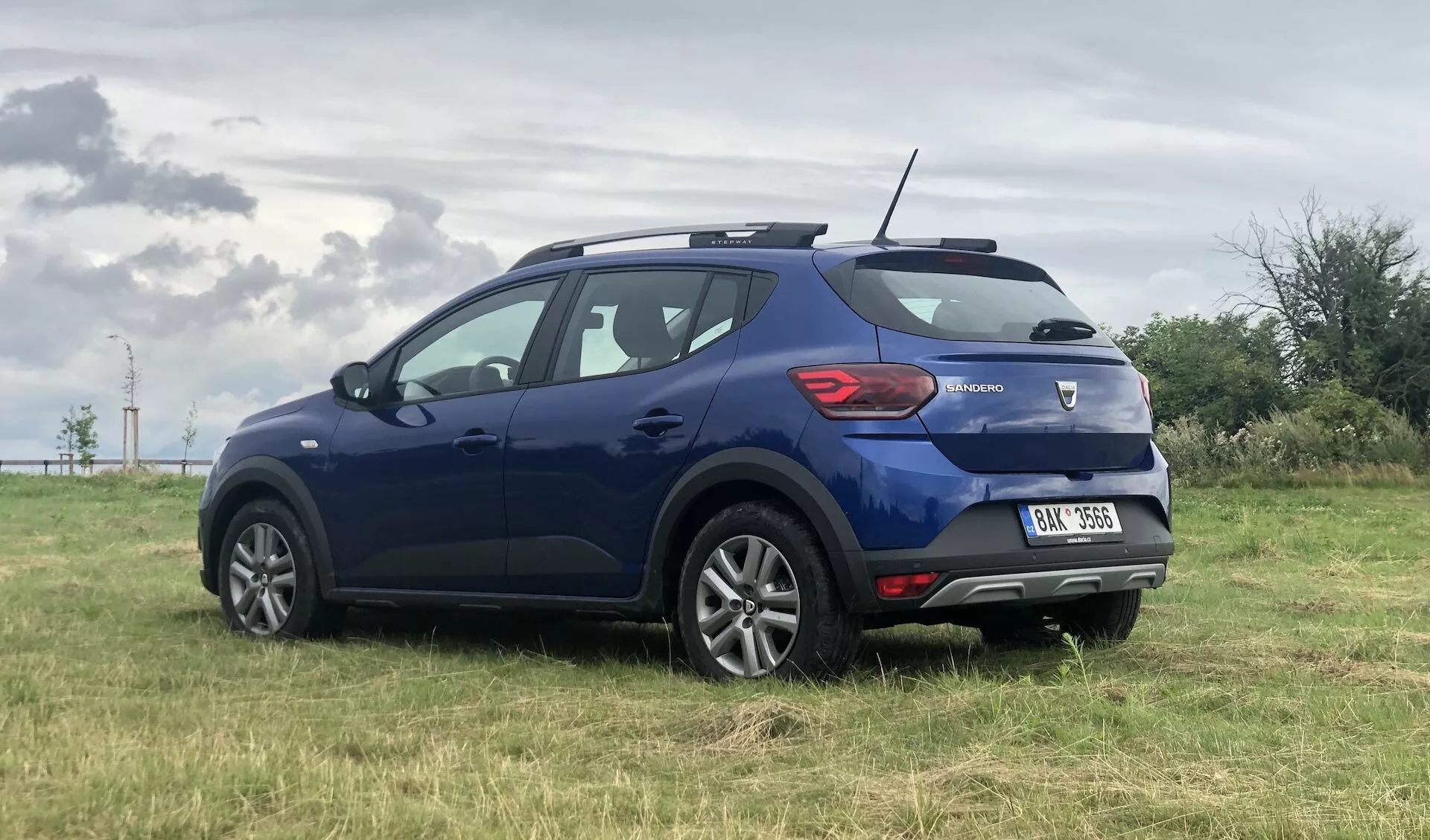 Trikotni zadnji pogled na modrenega kompaktnaga SUV Dacia Sandero Stepway 2020, parkiranega na travniku pod oblačnim nebom. Vidne lastnosti vključujejo hatchback karoserijo, zadnji spojler, zadnje luči, ki se ovijejo od zadaj do boka, lahka platišča, anteno v obliki plavuti morskega psa, zatemnjene zadnje steklo, znak nad zadnjo registrsko tablico, integriran reflektor in vzvratno luč v spodnjem zadnjem odbijaču ter stranska ogledala z integriranimi smerniki.