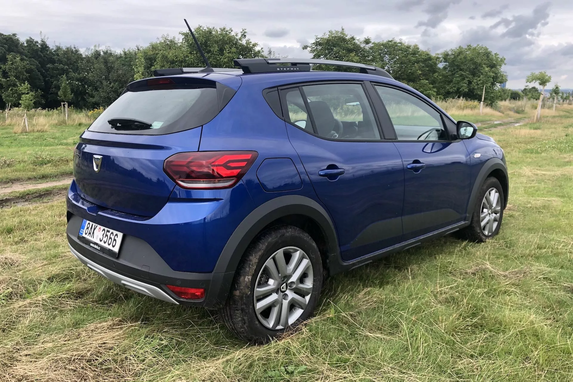 Zadnji tritretjinski pogled na sodoben moder crossover SUV Dacia Sandero Stepway 2020, parkiran na travnatem terenu, prikazuje eleganten dizajn, lita platišča, strešne nosilce, anteno v obliki plavuti morskega psa, zadnje kombinirane luči in zadnjo značko pod čistim, naravnim nebom.