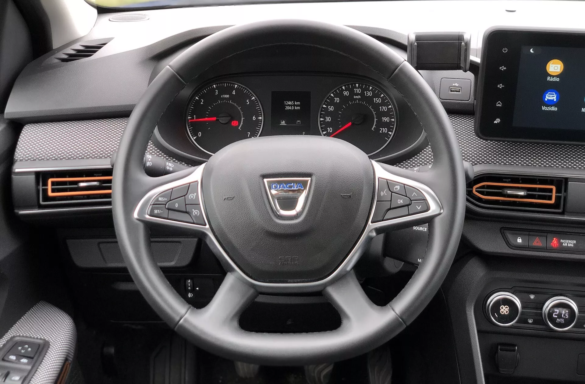 Notranji pogled na Dacia Sandero Stepway 2020 s poudarkom na voznikovi perspektivi armaturne plošče. Volan, postavljen v sredino fotografije, ima na sredi proizvajalčev logotip in več kontrolnih gumbov za funkcije, kot so upravljanje glasnosti, prostoročno klicanje in tempomat. Za volanom je viden instrumentni del z analognimi številčnicami za merilnik hitrosti in merilnik vrtljajev ter digitalni zaslon med njima. Na sredini armaturne plošče se nahaja zaslon na dotik z infotainment sistemom, ki prikazuje možnosti povezljivosti, kot je Bluetooth. Vidni so tudi zračniki z oranžnimi poudarki, dvo-conski klimatski nadzor in namigi o voznikovih vratih z električnim upravljanjem oken in nastavitvami stranskih ogledal.