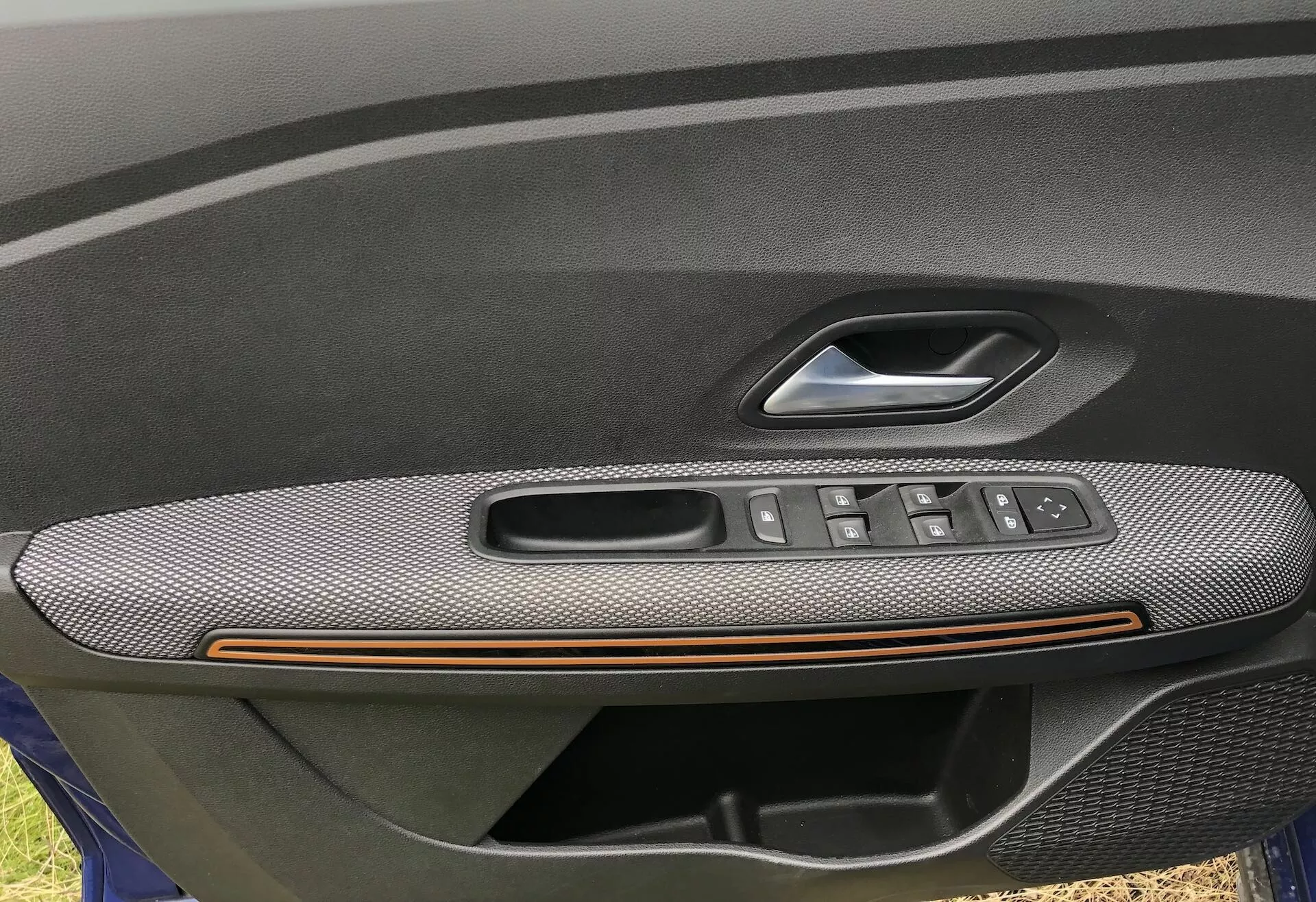 Posnetek od blizu notranje plošče vozniških vrat avtomobila Dacia Sandero 2020 Stepway, ki prikazuje naslon za roko z teksturo iz ogljikovih vlaken, bronasto poudarjeno linijo in vgrajeno nadzorno enoto s tipkami za električna okna, krmilno palčko za nastavitev stranskih ogledal in ročaj vrat z gumbom za zaklepanje/odklepanje. Sliko poudarjajo luksuzne in priročne funkcije, kar nakazuje višjo raven opreme.
