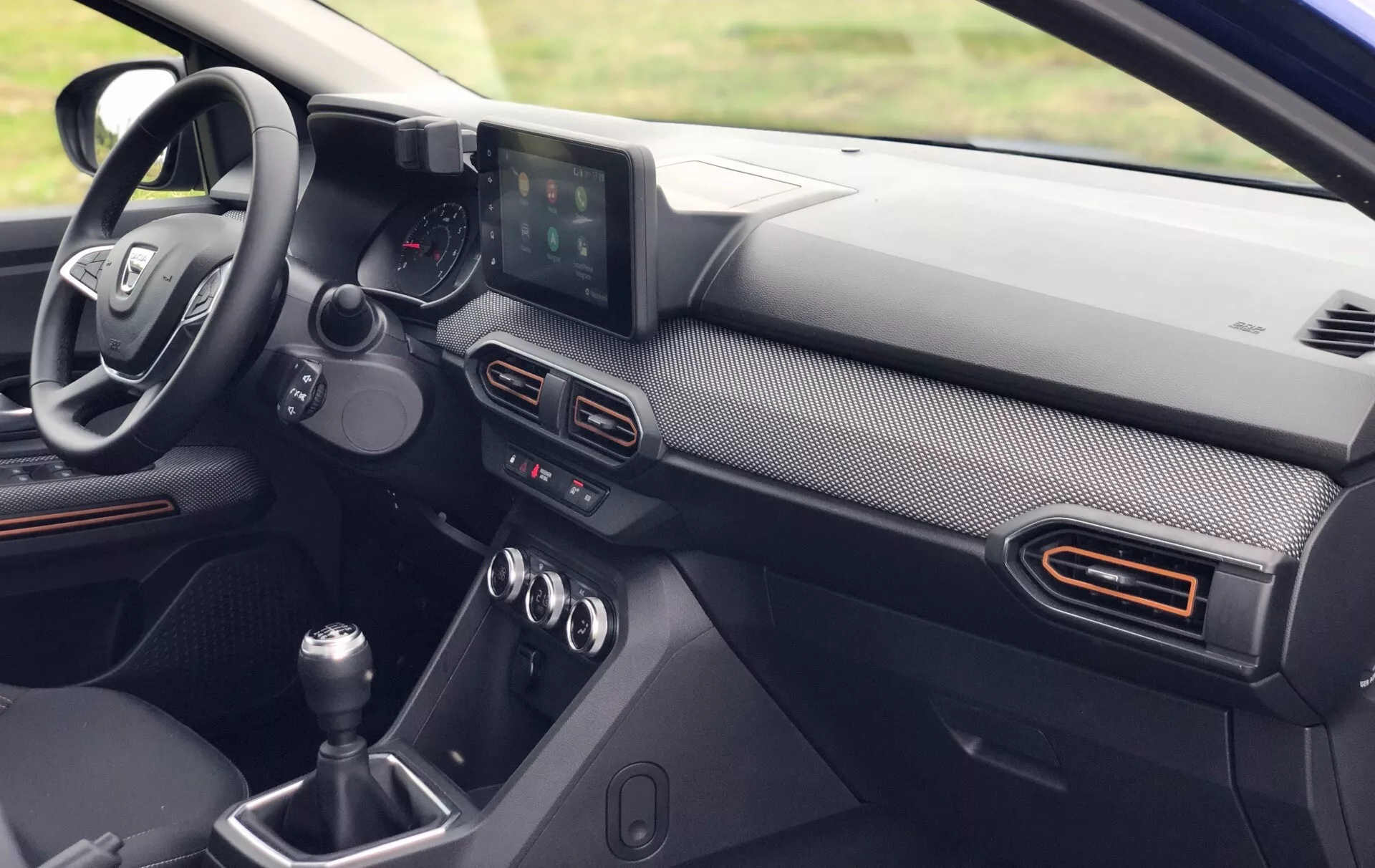 Pogled od blizu na notranjost Dacia Sandero Stepway 2020, ki prikazuje voznikovo stran armaturne plošče iz perspektive sprednjega potnika. Vidne funkcije vključujejo večfunkcijski volan s kontrolnimi gumbi, analogno in digitalno kombinirano instrumentno ploščo, samostoječ zaslon za informacijsko-zabavni sistem ter ročico ročnega menjalnika. Armaturna plošča ima oblogo, ki posnema ogljikova vlakna, in morebitne barvne poudarke okoli zračnikov, kar nakazuje na sodoben in morda zmogljivostno usmerjen dizajn.