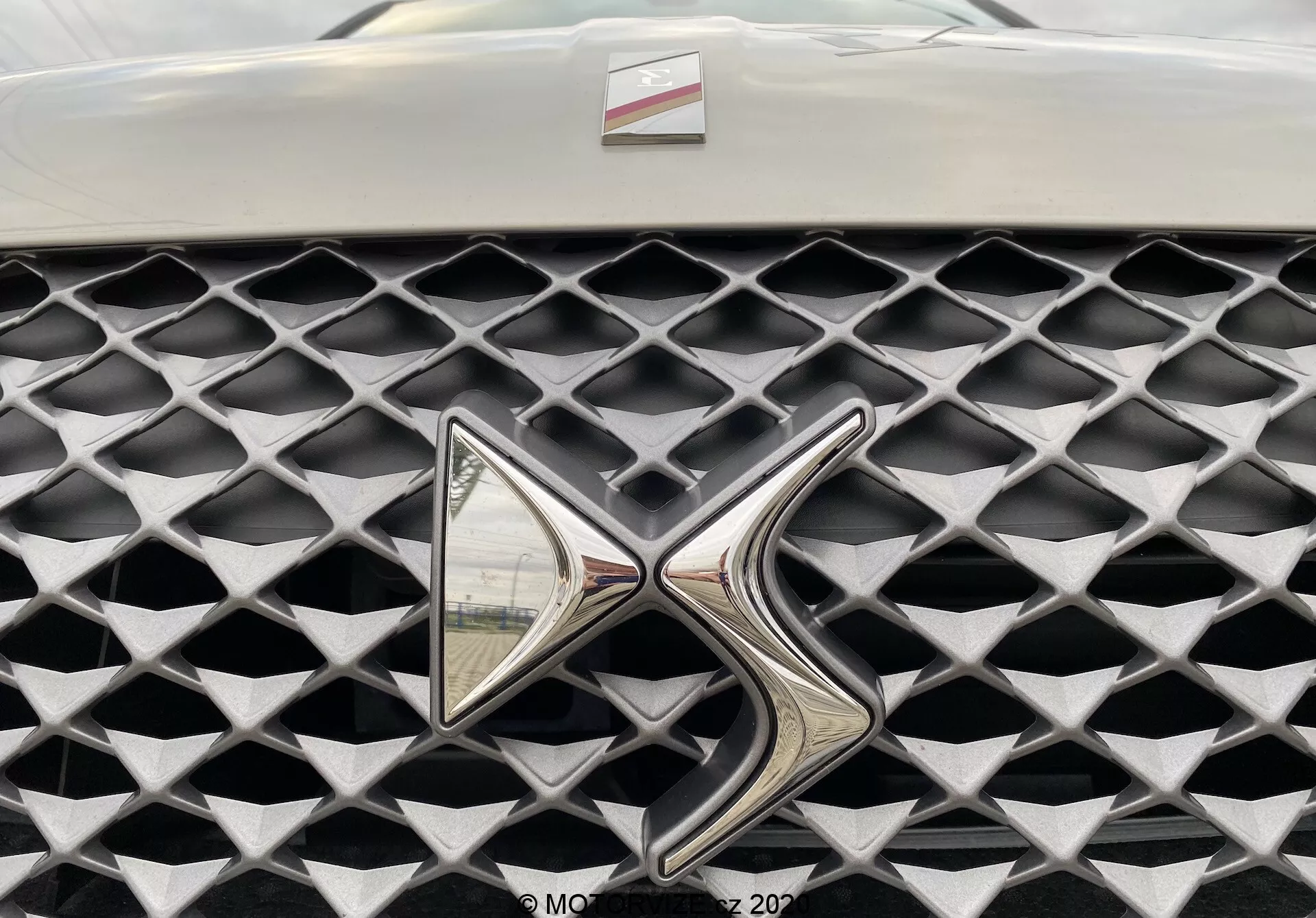 Povečava na masko modela DS Automobiles DS 3 Crossback E-Tense 2019. Slika zajema masko iz čelne perspektive, osredotočena na izstopajoči, močno odsevni kovinski znak. Maska ima značilno teksturo iz diamantnih celic v mat sivi barvi, kar kontrastira s sijajnim kromiranim znakom. Omejeno vidno polje onkraj maske in znaka, zamegljeno ozadje.