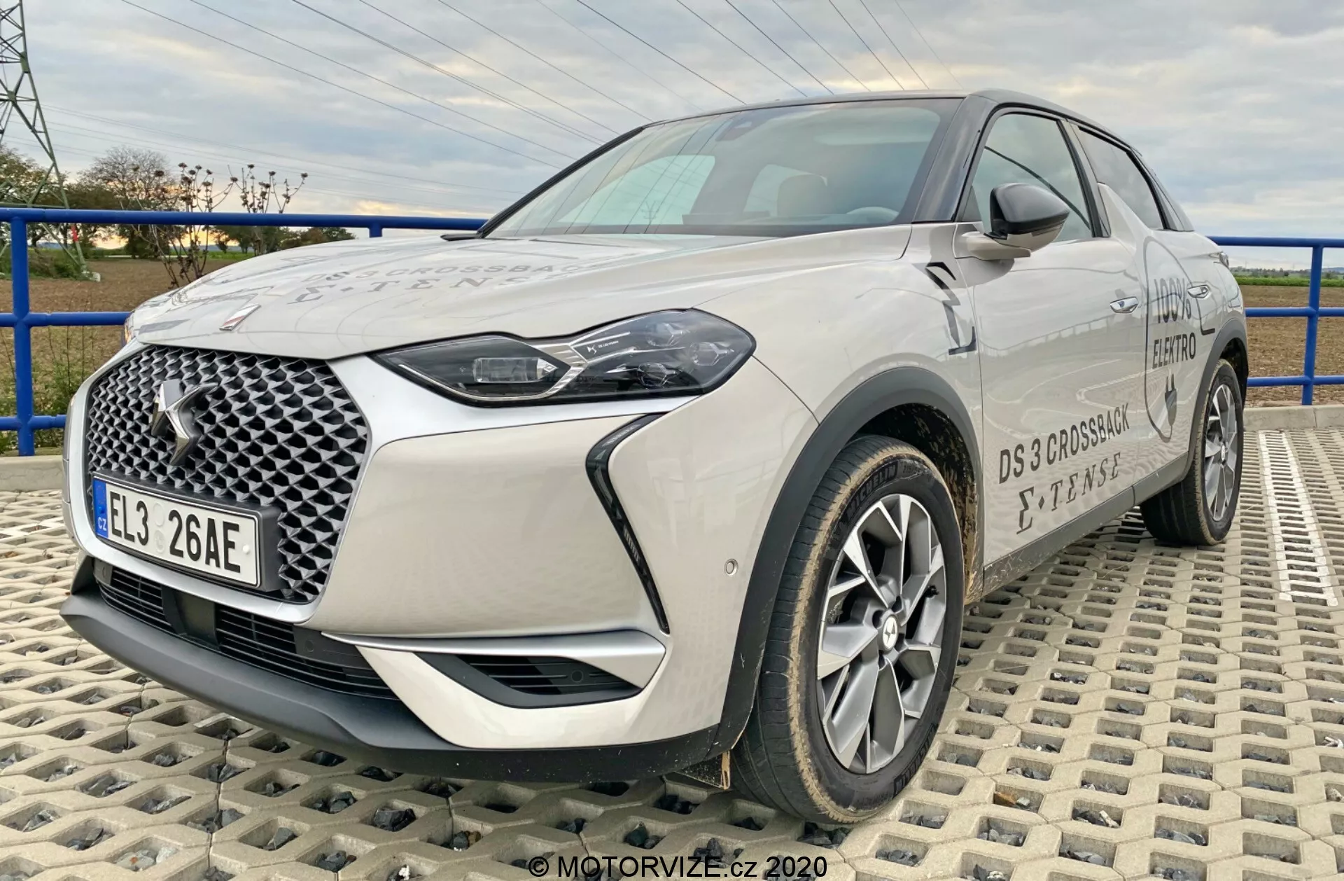 Slika prikazuje sodoben crossover SUV DS-Automobiles DS-3 2019 (DS-3 Crossback E-Tense), zajet iz kota treh četrtin spredaj na teksturirani površini, narejeni iz prepletajočih se ploščic. Perspektiva iz nizke do srednje višine poudarja status avtomobila in ponuja celovit pogled na njegovo sprednjo rešetko z značilnim geometrijskim vzorcem, LED žarometi, lita platišča, ogledala in podrobnosti blagovne znamke. Jasno vidna sta dolžina in sprednji dizajn vozila, kar poudarja njegovo sodobno in napredno avtomobilsko tehnologijo.