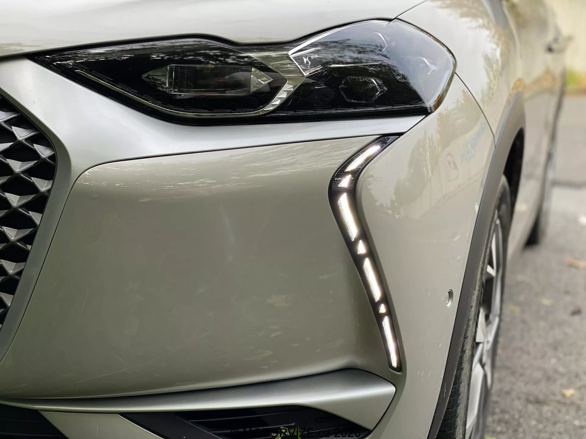 Pogled od blizu na sprednji levi kot modernega avtomobila DS Automobiles DS-3 Crossback E-Tense iz leta 2019. Slika se osredotoča na eleganten, kotni LED dizajn žarometov z LED dnevnimi lučmi vzdolž zgornjega roba in del mreže z diamantnim vzorcem, poudarja luksuz in vrhunsko tehnologijo.