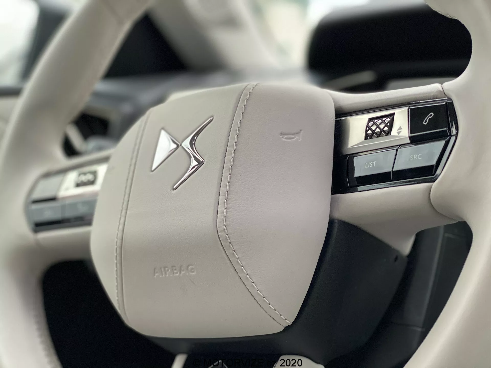 Bližnji posnetek leve strani modernega, svetlega usnjenega volana avtomobila DS Automobiles DS 3 Crossback E-Tense 2019, ki poudarja izdelavo in razkošje z vidnimi šivi. Vključuje več integriranih gumbov za nadzor funkcij vozila, vključno z glasovnimi ukazi, telefonom, glavnim menijem in upravljanjem medijev, poleg izrazitega osrednjega emblema in ohišja z napisom 'AIRBAG'.