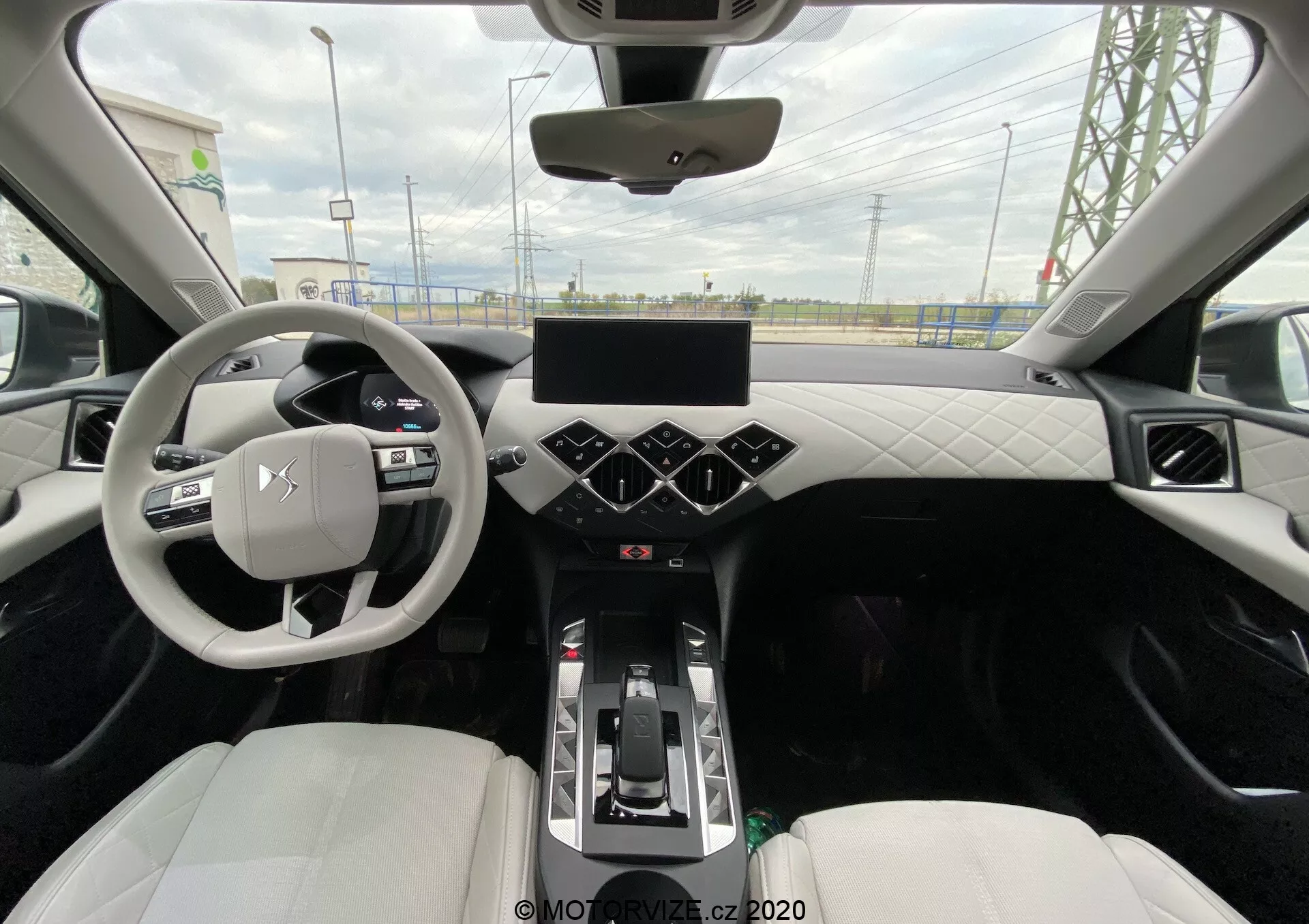 Pogled s zadnjega sedeža na moderen centralni notranji prostor vozila DS Automobiles DS-3 Crossback E-Tense 2019. Fotografija zajema volan s tremi kraki z nameščenimi kontrolnimi gumbi, digitalni prikazovalnik instrumentov, velik zaslon na dotik na sredinski konzoli s heksagonalno razporejenimi gumbi za klimatsko napravo pod njim ter prestavno ročico, kar nakazuje avtomatski menjalnik. Armaturna plošča in vrata imajo prešite vzorce iz usnja ali usnje podobnega materiala, sprednji športni sedeži so oblazinjeni v svetlo usnje ali sintetični material. Vidne so ambientalne luči vzdolž obrobe, panoramska streha in zračne reže na armaturni plošči.