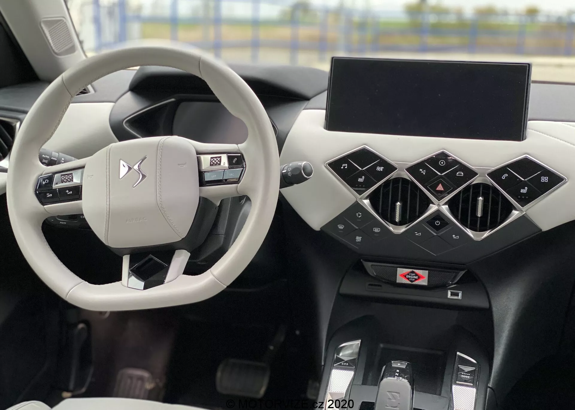 Notranji posnetek DS Automobiles DS 3 Crossback E-Tense 2019 s poudarkom na sredinsko postavljenem srebrnem/svetlosivem volanu z upravljalnimi gumbi in logotipom proizvajalca, skozi katerega je viden voznikov sedež. Črna armaturna plošča ima geometrijske elemente s trikotnimi sredinskimi zračniki, izklopljen zaslon infotainmenta, spodaj pa verjetno kontrole klimatske naprave in osrednji tunel. Na levi je delno viden voznikov vrat z upravljanjem oken in ogledal.