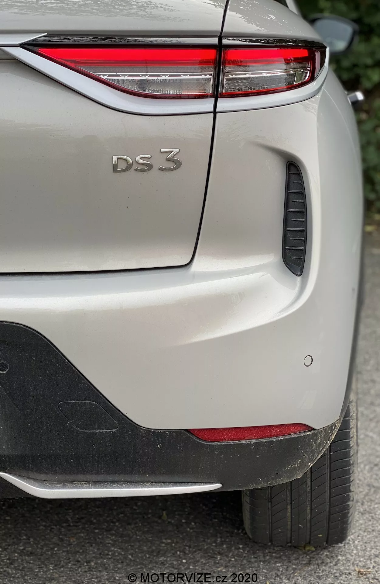 Pripoveden posnetek zadnjega stranskega pogleda DS Automobiles DS 3 Crossback E-Tense 2019, osredotočen na zgornji desni del zadnjega odbijača, pokrov prtljažnika in del zadnjega desnega blatnika. Slika zajema značilne oblikovalske podrobnosti modela, vključno z ozkimi zadnjimi LED lučmi, reliefnim napisom 'DS 3', parkirnimi senzorji in črnim plastičnim elementom na odbijaču. Avto, prikazano v svetlem odtenku z metalnim zaključkom, je fotografirano v naravni svetlobi, kar poudarja drobne podrobnosti laka in teksture.