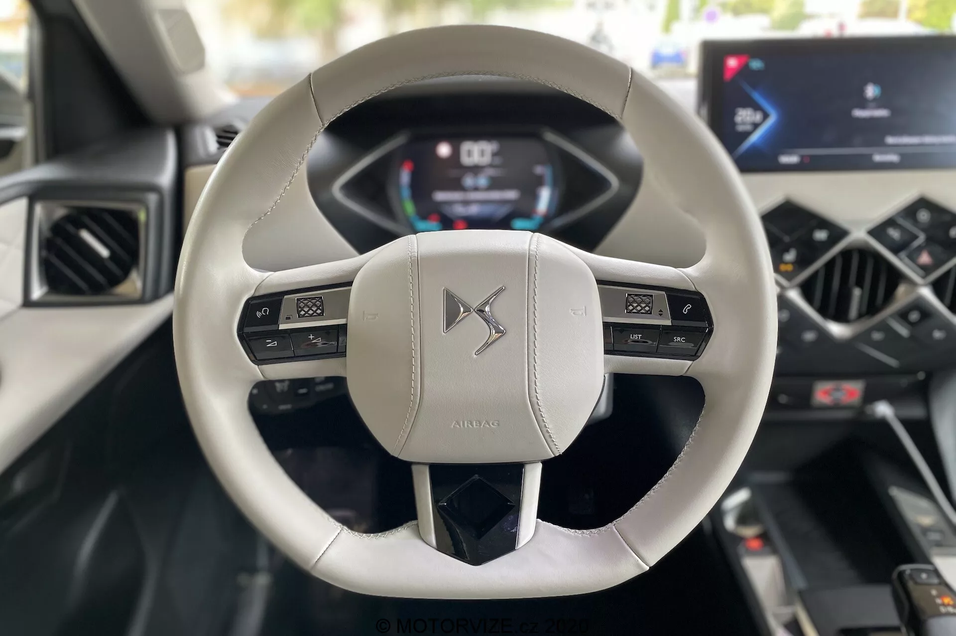 Notranji pogled z perspektive voznika v DS Automobiles DS 3 Crossback E-Tense 2019, s poudarkom na modernem dvokrakem volanu, oblečenem v svetlem usnju, z večfunkcijskimi gumbi in digitalno instrumentno ploščo, ki prikazuje hitrost in vrtljaje. Osrednja konzola se ponaša z integriranim zaslonom na dotik za informacije in zabavo ter minimalističnimi kontrolami klimatske naprave, kar poudarja napredno avtomobilsko tehnologijo in luksuzno oblikovanje.